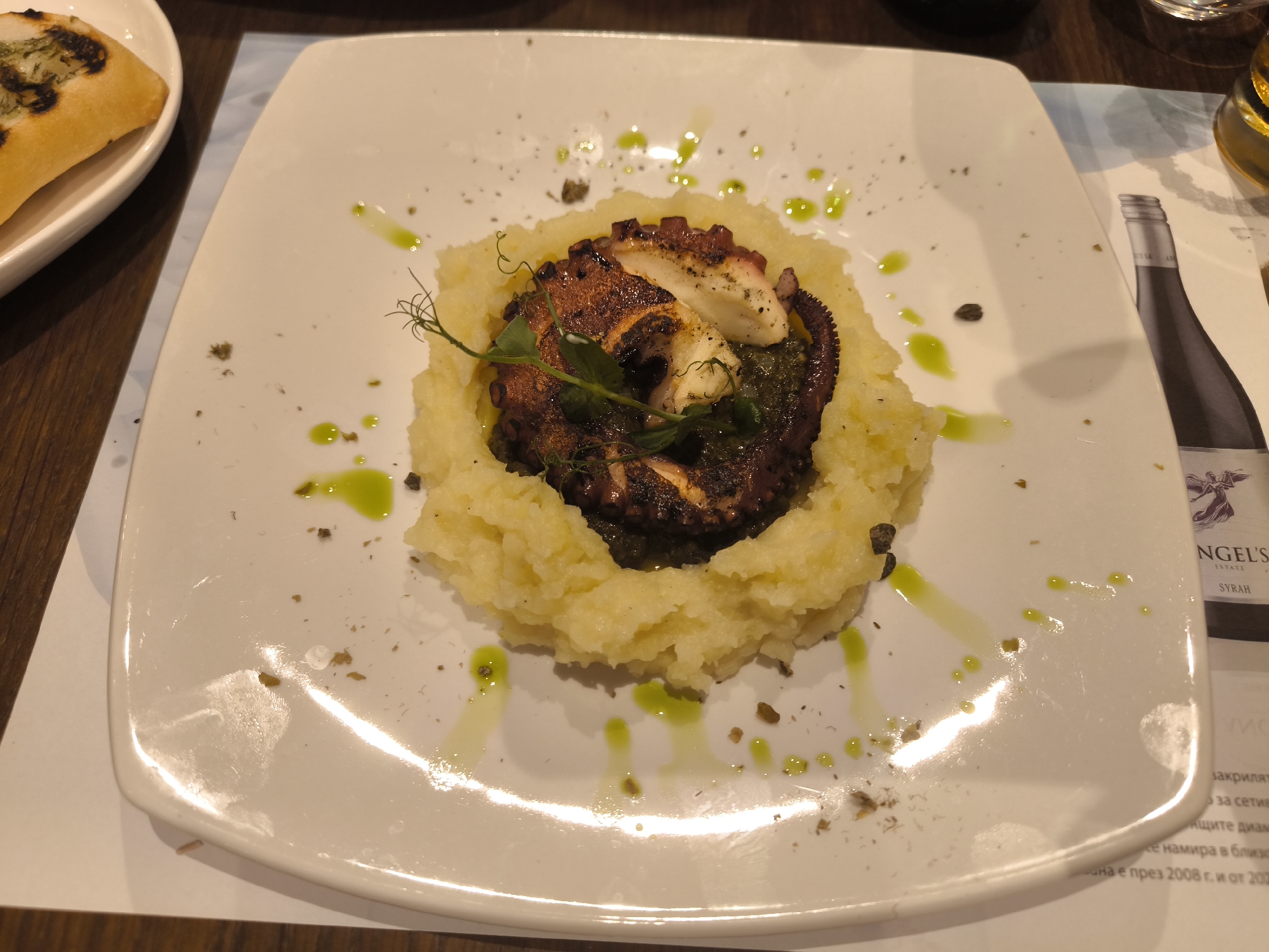 The image shows a plate of food, likely in a restaurant. The main dish appears to be grilled octopus served on a bed of mashed potatoes and a green sauce. The plate is white and square with rounded edges. The food is drizzled with a green oil or sauce, and there are some small dark specks scattered around the plate. To the left, a partially visible bread item rests on a separate plate. To the right, there is a bottle of wine and some printed material. The scene takes place at a table in Unknown, Unknown.