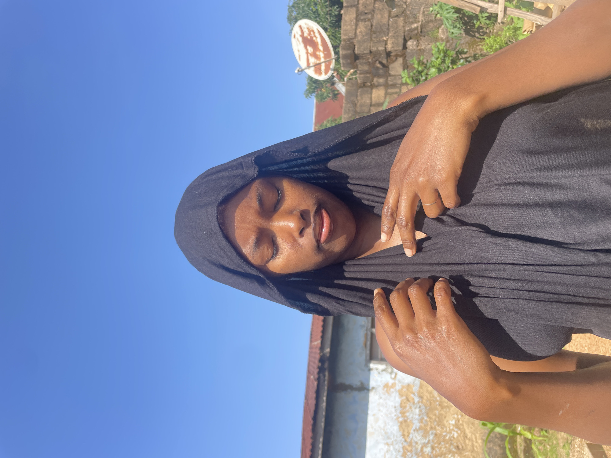 A medium shot captures a young Black woman outdoors, her eyes closed and head tilted slightly down, as she is wrapped in a black headscarf. Her hands are raised and gently placed near her chest, with her fingers interlocked in the left hand. The background features a clear blue sky and hints of a residential area in Jos, Nigeria, with a weathered building with a corrugated metal roof on the left, and a brick wall and satellite dish on the right. The lighting suggests a bright, sunny day.