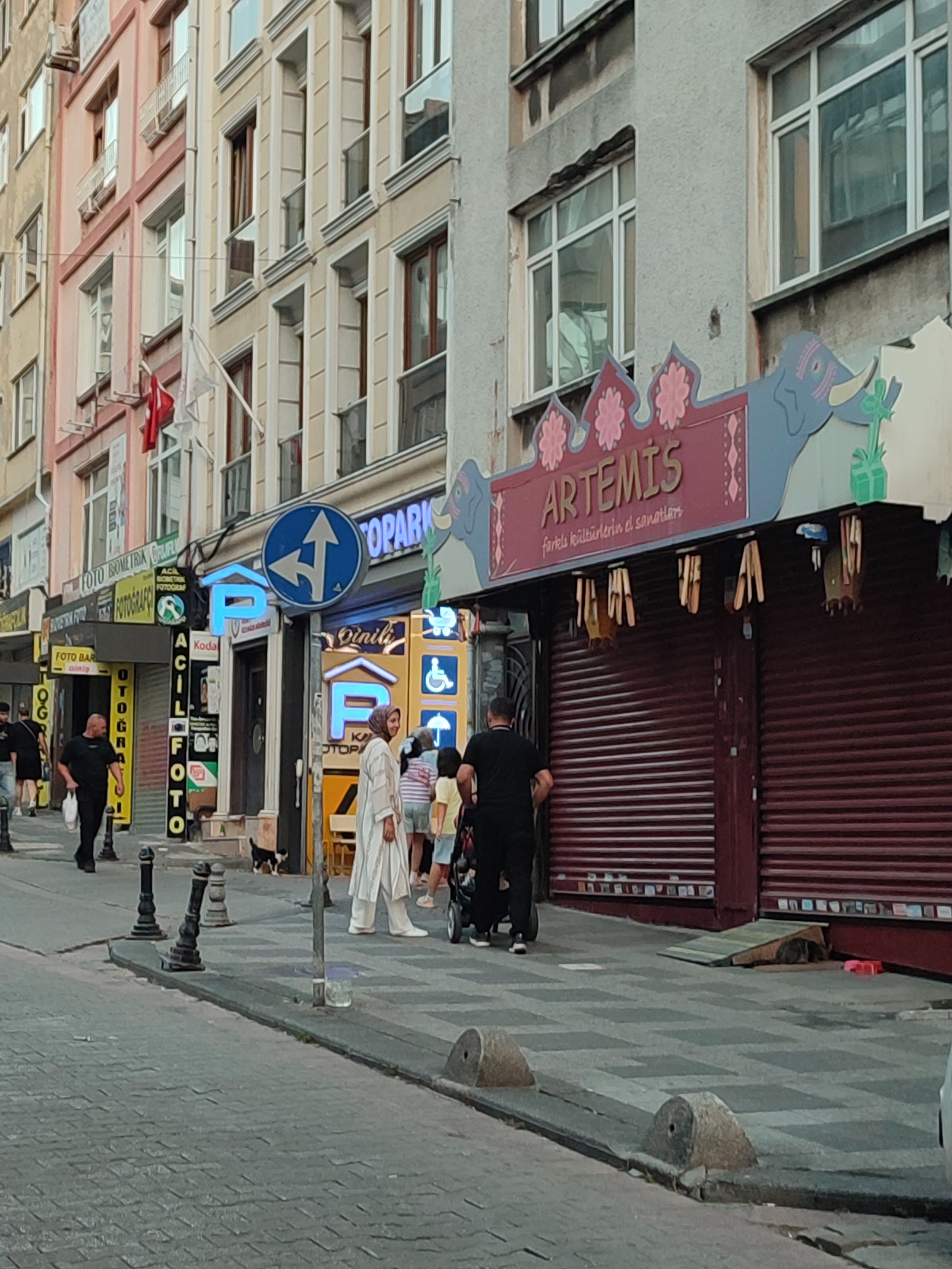 The image depicts a street scene in Kadıköy, Turkey, likely a commercial area. On the right side of the frame, a building displays a sign that reads "ARTEMIS" in large lettering, with smaller text beneath it that appears to be in Turkish. The storefront below the sign has dark red metal roll-down shutters. Several wooden objects, possibly decorative or functional items, are hanging from the overhang of the Artemis shop. To the left of the Artemis shop, a building has multiple signs, including one with a blue parking symbol and arrows, indicating traffic direction. Further left, another building features various storefronts with signs such as "FOTO ISOMETRIK", "FOTOĞRAFÇI", "FOTO BAR", and "ACIL FOTO".
In the foreground, people are walking along the sidewalk. A man in a black t-shirt and black pants is walking towards the left of the frame, carrying a white bag. Further down the street, another man in black attire is visible. A woman in a long, white, patterned robe is walking towards the right, with a few other individuals following her, including a child in a striped shirt. A stroller is also present. On the sidewalk, a small black and white cat is walking. The street is paved with cobblestones and has bollards lining the edge of the sidewalk. The architecture suggests a mix of older and newer buildings, typical of urban environments. The overall atmosphere appears to be that of a busy street during daylight hours.