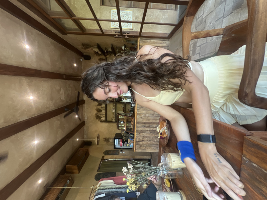 A woman with curly brown hair sits at a wooden table in a cafe with exposed beams and a vintage interior. She wears a light yellow top and skirt. Her left wrist has a blue band, and her right wrist has a fitness tracker and a small tattoo. A vase of flowers and drinks sit on the table. A bakery display case is visible in the background.