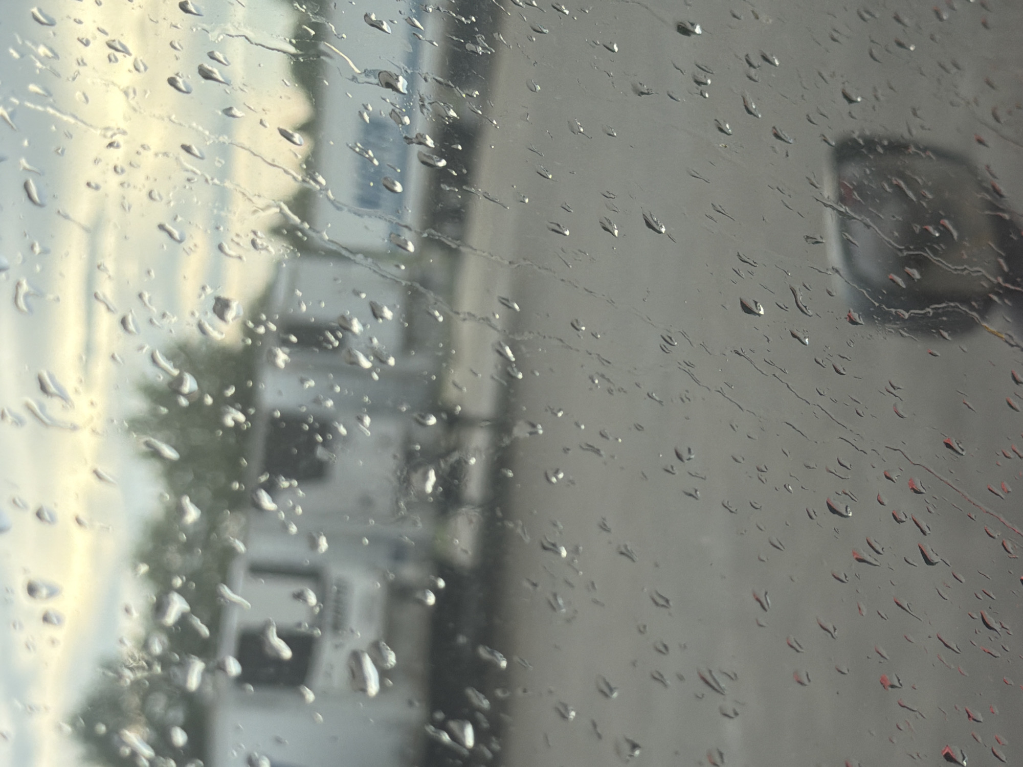 The image shows a view through a rain-streaked window, likely from inside a vehicle. In the background, several white semi-trailers are parked, suggesting a commercial or industrial area. The sky above appears overcast with hints of pale yellow light, possibly indicating dawn or dusk. The foreground is dominated by numerous raindrops on the glass, distorting the view of the scene beyond. The ground appears to be a paved surface, possibly asphalt. A blurred reflection of what looks like a vehicle's side mirror can be seen in the lower right portion of the image. The presence of semi-trailers suggests the location could be a truck stop, distribution center, or a similar facility in or around Murfreesboro, Tennessee. There are no clearly discernible people or specific actions taking place.
