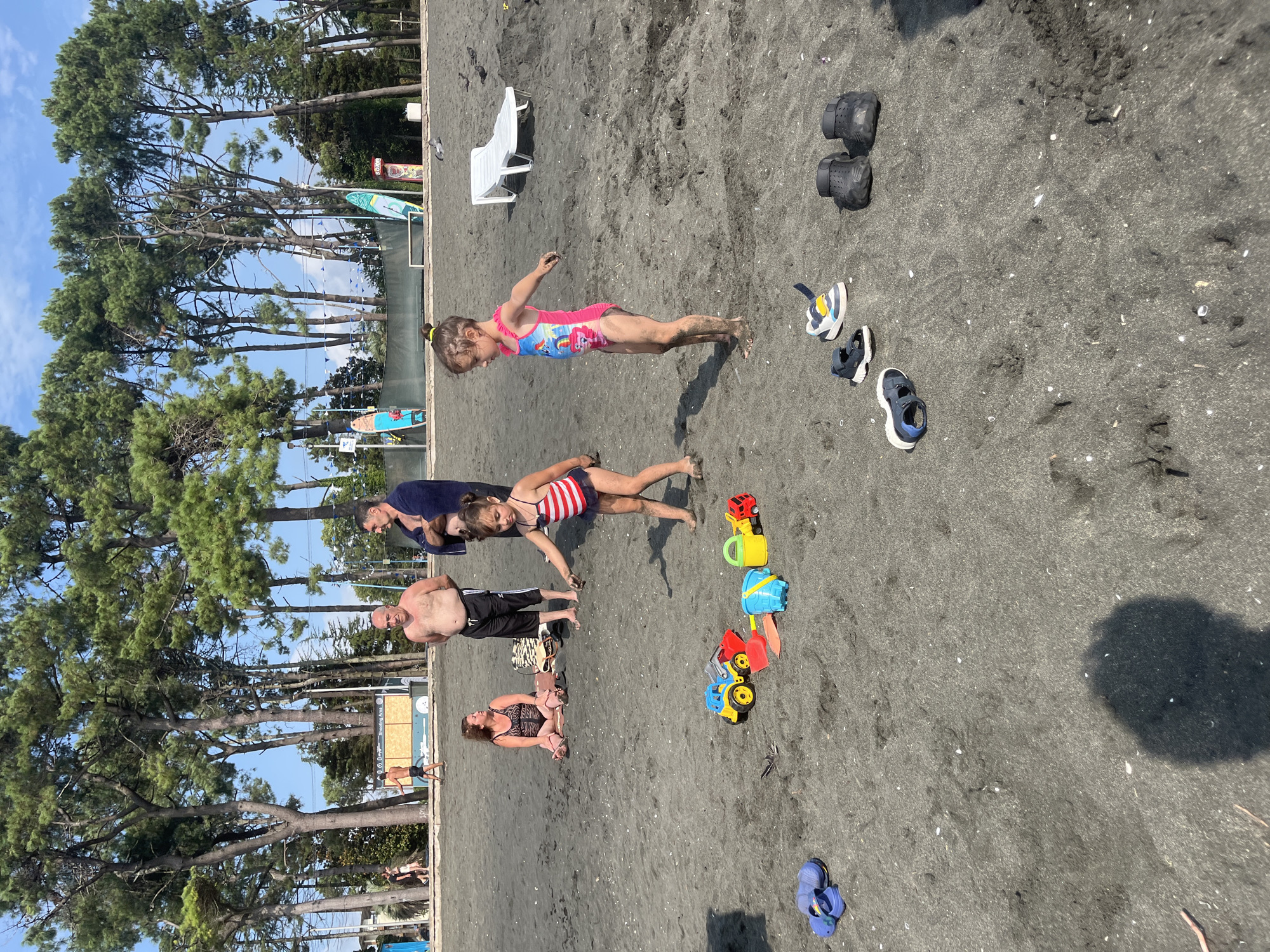 In Ureki, Georgia, on a black sand beach, two young girls play with toy vehicles and buckets in the shallow water. One girl, wearing a red and white striped swimsuit, is actively engaged with the toys, while the other, in a pink swimsuit, is also playing. Two adults, likely parents, stand nearby observing. A man with a towel draped over his shoulder and another man without a shirt are present. In the background, a row of tall pine trees stands against a bright blue sky, and a white lounge chair is visible. The scene suggests a relaxed family outing at the beach.