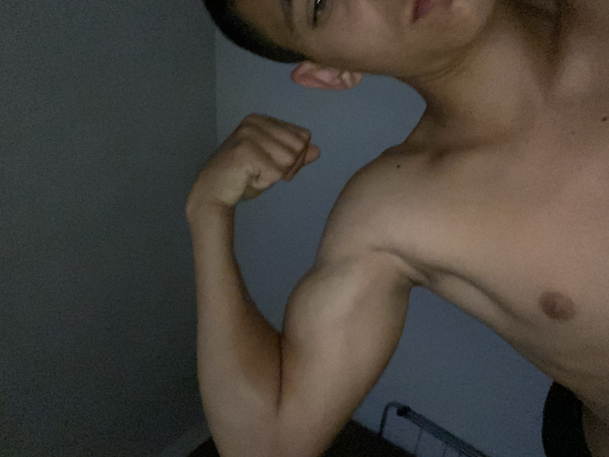 A close-up, low-light, waist-up shot of a young person flexing their bicep. The subject is positioned in the center of the frame, facing towards the right side of the image. The background is a dark, textured wall, with a faint blueish hue. The lighting is dim, creating soft shadows on the subject's skin, highlighting the muscles of the arm. The person has short dark hair and is looking towards the camera with a slight smile. There are no discernible objects or locations in the image that would allow for a specific geographical placement within Brashlen, Bulgaria.