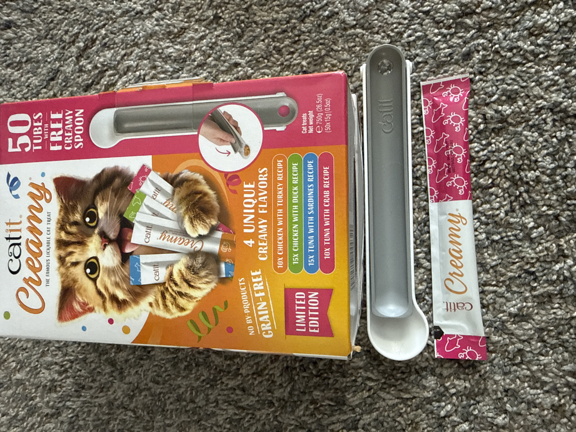 An overhead shot displays a box of Catit Creamy cat treats, a cat treat tube, and a grey and white cat treat dispenser, all situated on a grey carpeted floor. The box is predominantly orange and white and features an image of a kitten. Text on the box reads "Catit Creamy," "The Famous Lickable Cat Treat," "50 Tubes," "With Free Creamy Spoon," "No By-Products," "Grain-Free," "4 Unique Creamy Flavors," and a list of flavors with quantities: "10x Chicken with Turkey Recipe," "15x Chicken with Duck Recipe," "15x Tuna with Sardines Recipe," and "10x Tuna with Crab Recipe." A pink banner on the box indicates "Limited Edition." The treat tube is white with pink crabs and fish, and the text "Catit. Creamy." The dispenser is grey and white with the "Catit" logo. The image was taken in Girard, United States of America.