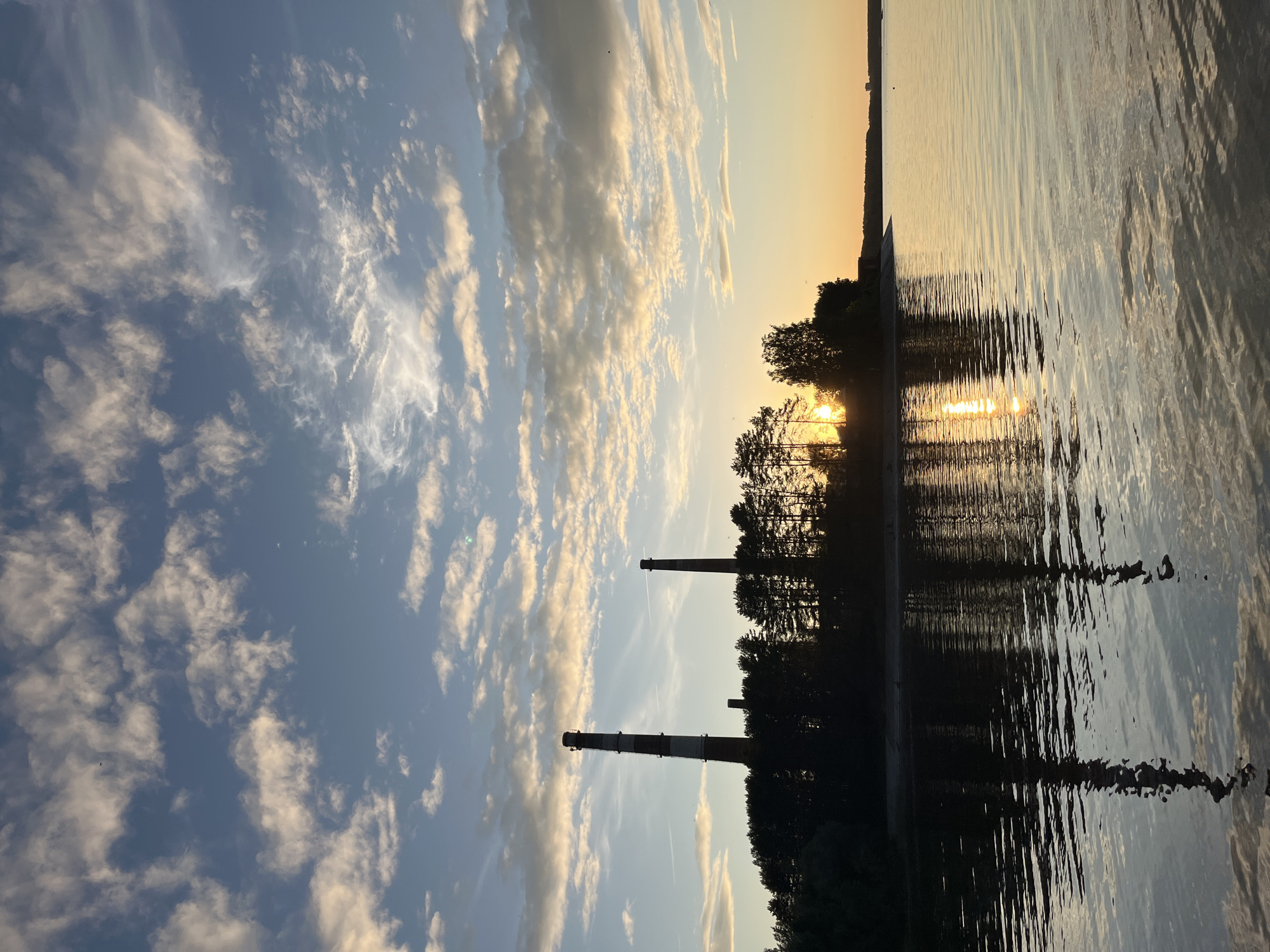 This image depicts a sunset scene over a body of water in Chaikovsky, Russia. The sky, predominantly blue, is filled with scattered, illuminated cumulus and cirrus clouds, exhibiting warm golden and grey tones, with subtle contrails visible in the upper sections. The horizon reveals a silhouetted landmass, featuring a dense line of trees and three industrial chimneys. The leftmost chimney is significantly wider and taller, displaying red and white banding, while the central and rightmost chimneys are narrower and darker. The setting sun, a bright golden orb, is partially obscured by trees on the right side of the frame, casting an intense golden glow across the water. The calm surface of the water perfectly mirrors the sky's colors, the sun's reflection, and the silhouettes of the chimneys and trees, creating elongated, shimmering vertical reflections. No discernible human figures or immediate human activity are present.