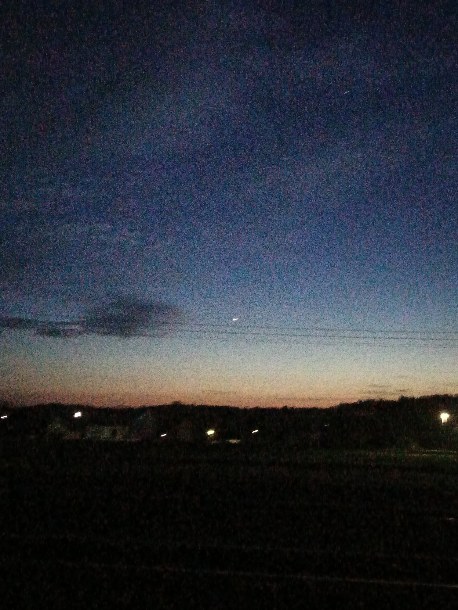The image depicts a landscape at dusk or night, possibly located near or within a city. The sky transitions from dark blue at the top to orange near the horizon. A dark cloud is visible in the middle-left of the frame. In the distance, lights suggest the presence of buildings or streetlights. A dark field occupies the foreground. The location is possibly in or near Лосева, Belarus.