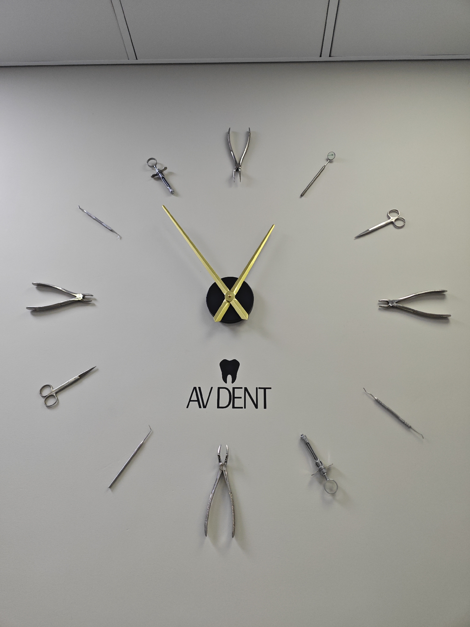This is a full, eye-level, medium shot of a wall clock with a minimalist design. The clock face is the wall itself, and the numbers are represented by various dental instruments. The clock hands are gold and extend from a black circular center. The hours are marked by:
* **12 o'clock:** A pair of dental forceps.
* **1 o'clock:** A dental mirror.
* **2 o'clock:** A pair of surgical scissors.
* **3 o'clock:** A pair of dental pliers.
* **4 o'clock:** A dental probe.
* **5 o'clock:** A syringe.
* **6 o'clock:** A pair of dental extractors.
* **7 o'clock:** Another dental probe.
* **8 o'clock:** A pair of surgical scissors.
* **9 o'clock:** A pair of dental pliers.
* **10 o'clock:** A dental scaler.
* **11 o'clock:** A pair of dental forceps.
Below the clock's center is a black logo consisting of a stylized tooth and the text "AV DENT". The wall is a neutral gray color, and the lighting is even, suggesting an indoor environment. The image does not contain any people, actions, or interactions. There is no information in the image to indicate the location in the city of Lobnya, Russia.
