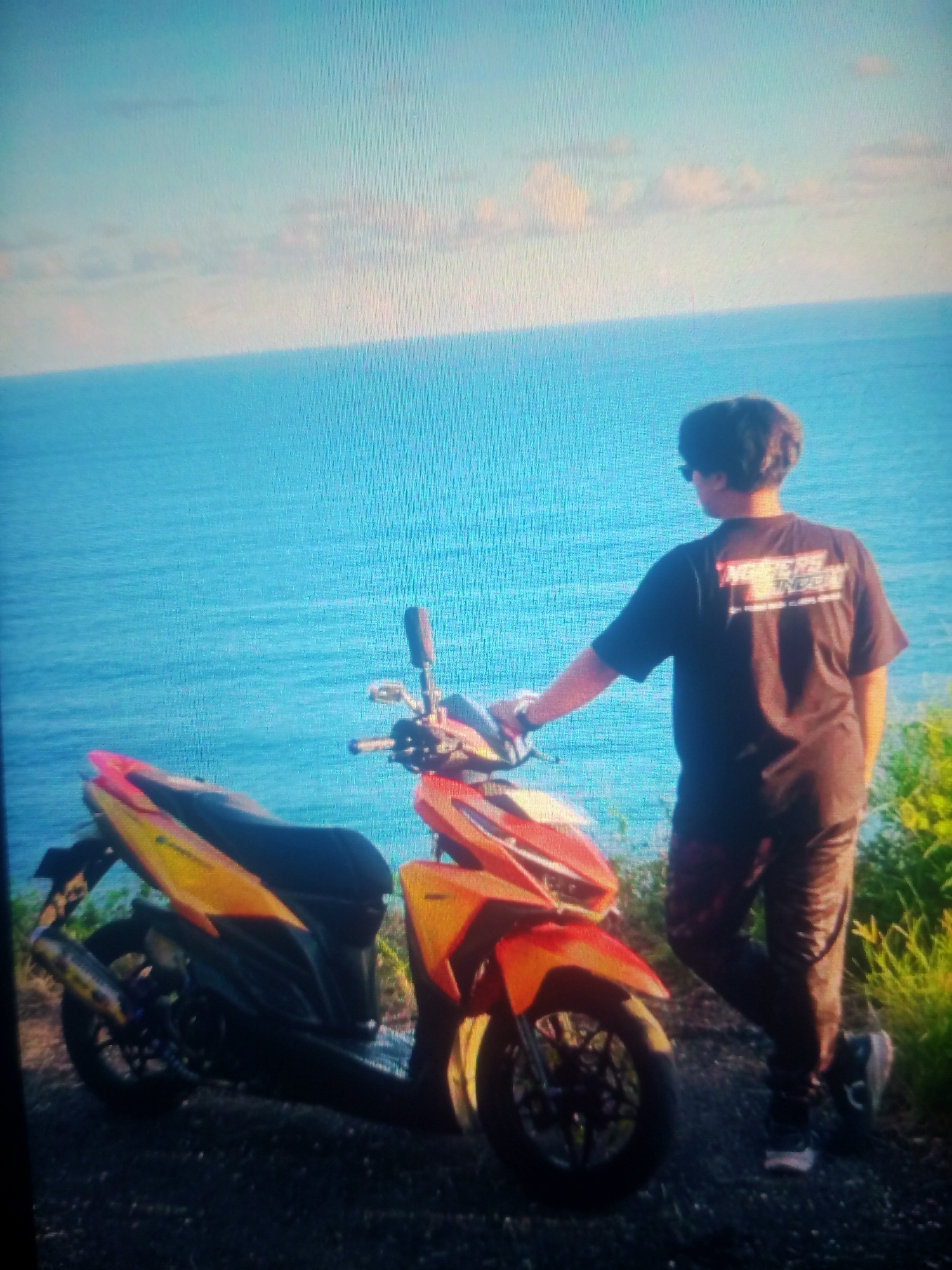 A person stands next to a motorcycle by the ocean. The person is facing away from the camera, wearing a black t-shirt and dark pants. The motorcycle is orange and yellow. The background is a bright blue ocean with some clouds. The scene is set in Nusa Agung, Indonesia.