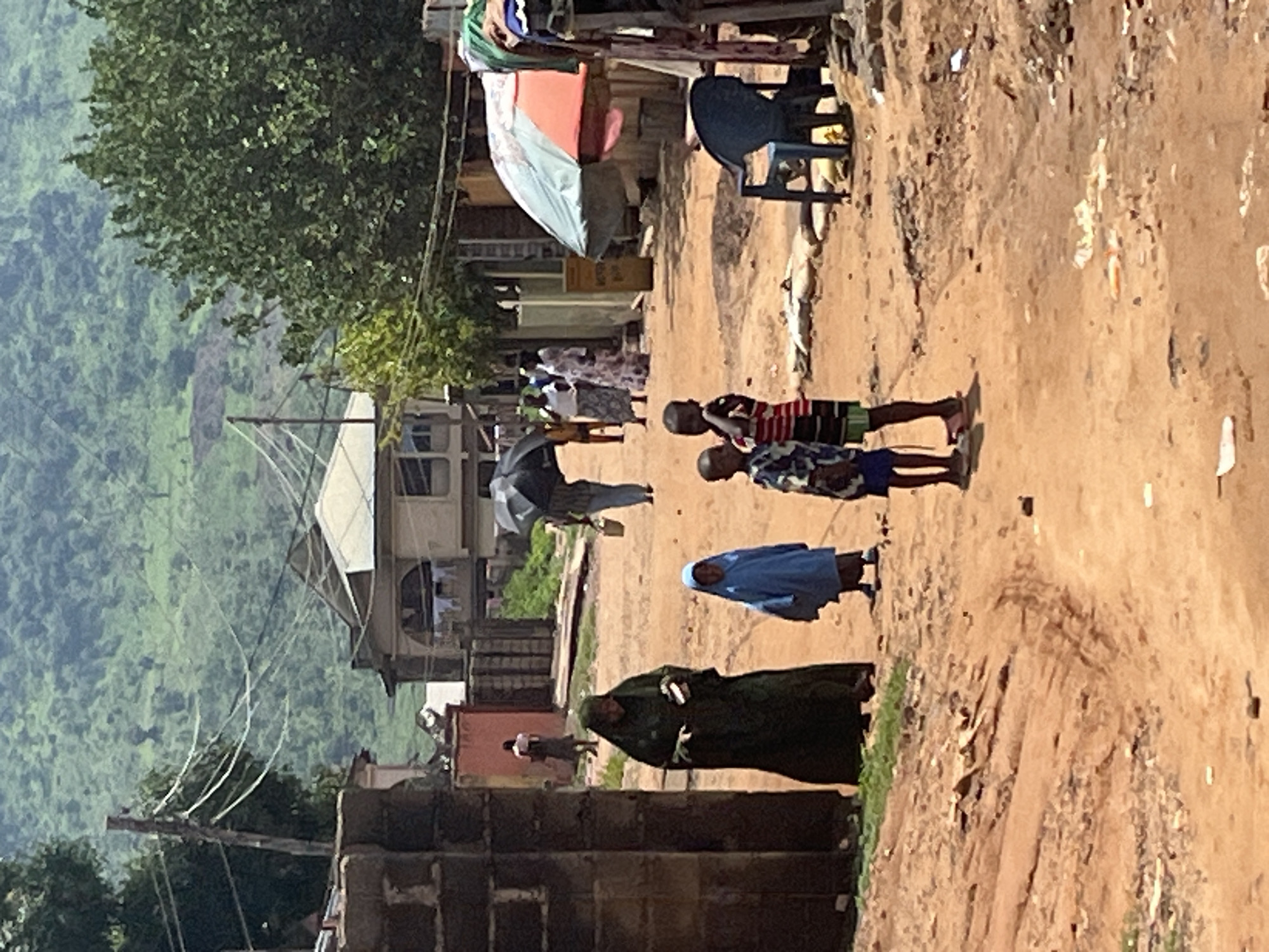 The image captures a street scene in a village with a dusty dirt road. In the foreground, two young boys stand on the right, one wearing a striped shirt and shorts, the other with a shirt and shorts. To their left, a young child in a blue robe stands on the road. Further left, a woman in a dark green robe is walking, holding something in her hand. In the background, several people are walking, with one person holding an umbrella and another carrying a bucket. Buildings with tin roofs line the street, and lush green hills are visible in the distance. There is a blue plastic chair to the right of the frame.