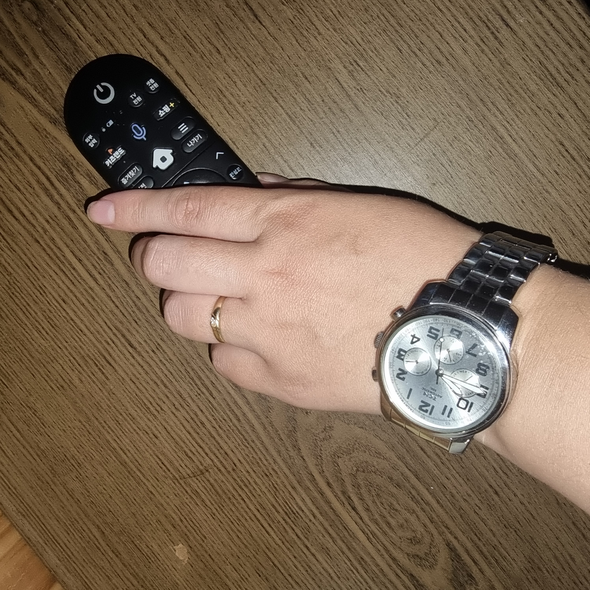 A person's left hand, adorned with a silver watch and a gold ring, rests on a wooden surface, holding a black remote control. The watch is a chronograph with a silver dial and a metal band. The remote control has several buttons, some with Korean characters. The wooden surface appears to be a table or desk, with a prominent wood grain pattern. The lighting is dim, creating shadows on the hand and the remote. No specific location in Dunnae-myeon, South Korea is discernible from the image.