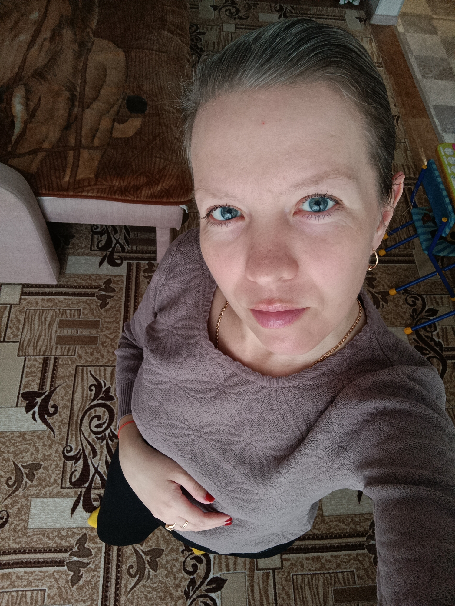A woman stands indoors in Chaikovsky, Russia, likely in a residential setting. The floor is covered with brown and beige patterned linoleum. A bed with a brown blanket depicting a wildlife scene is visible in the upper left. A small, colorful plastic chair is partially visible in the upper right.
The woman, viewed from a high angle, is wearing a mauve knitted sweater, a gold necklace, and gold hoop earrings. She has fair skin, blue eyes, and her hair is graying. She has rings on her fingers and red nail polish. Her left hand rests on her abdomen. She is looking directly at the camera.
