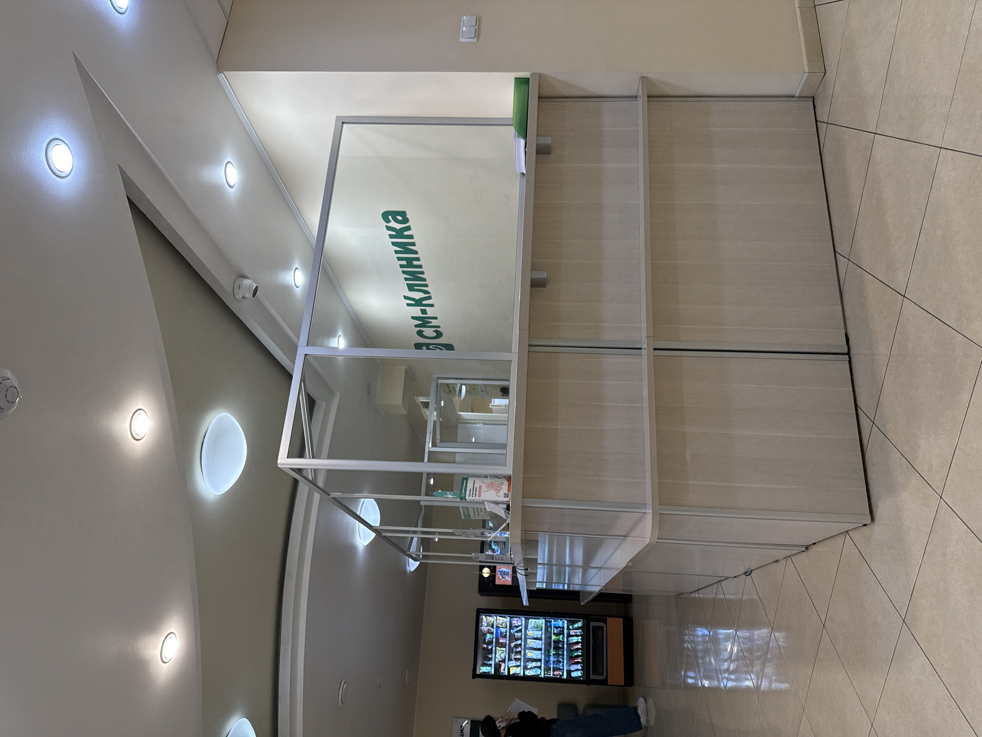 The image captures the interior of what appears to be a modern clinic or medical facility's reception or waiting area, seen from a high angle looking down and across the room. The space is well-lit with various ceiling fixtures, including circular recessed lights and larger oval-shaped ambient lights. The ceiling also features architectural details with sharp angles and a security camera.
In the foreground and central to the image is a sleek reception desk or counter made of glass and light-colored metal framing, featuring transparent partitions and shelves. Behind this structure are light wood-paneled cabinets or walls. Reflected on one of the glass panels is the green text "СМ-Клиника" (SM-Klinika), indicating a medical institution in a Russian-speaking country.
To the right side of the frame, the floor is covered in large, light-colored square tiles. Below the counter and near the right edge of the image, a person is partially visible from the waist down, wearing dark clothing and blue jeans, standing near a vending machine. The vending machine is well-stocked with various beverages and snacks, illuminated from within. The person appears to be holding a document or phone, possibly waiting or making a selection.
The overall setting suggests a clean, professional, and well-maintained environment. There are no clear indications of the time of day, but the interior is brightly lit by artificial lights.