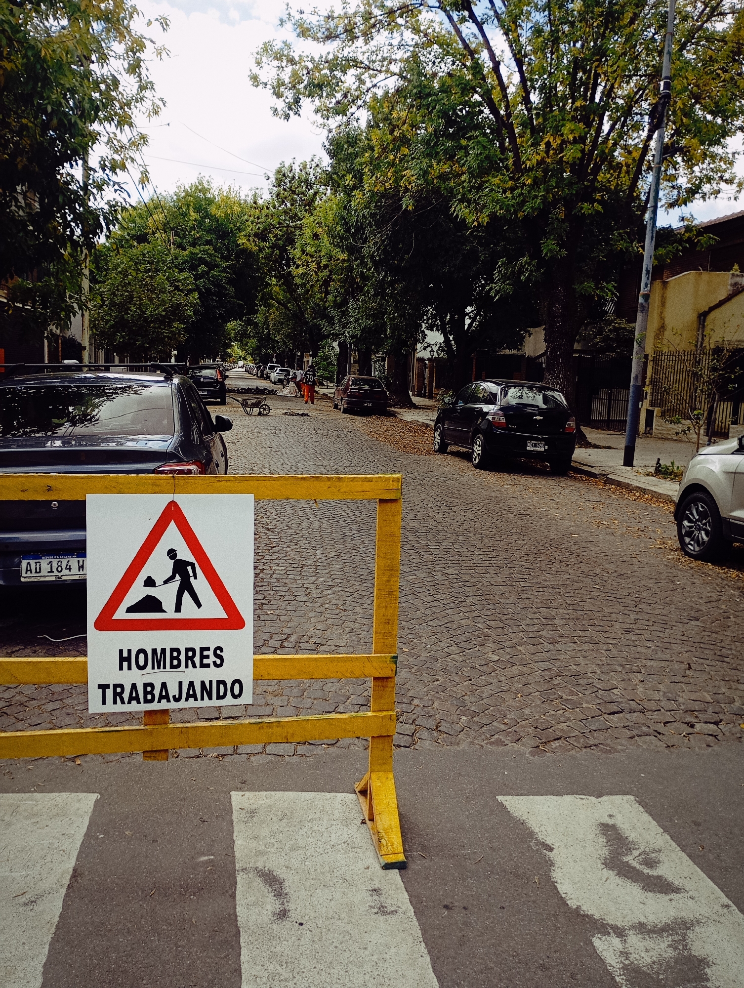 The image depicts a street scene in Buenos Aires, Argentina. The foreground shows a yellow barrier with a "Men at Work" sign ("Hombres Trabajando") in Spanish. A zebra crossing is visible beneath the barrier. Behind the barrier, a dark-colored car is parked. Further down the street, there are additional parked cars and trees lining both sides of the road. Two construction workers in orange vests are seen near a wheelbarrow. The road is paved with cobblestones. The sky is overcast. The license plate of the car in the foreground reads "AD 184 W".