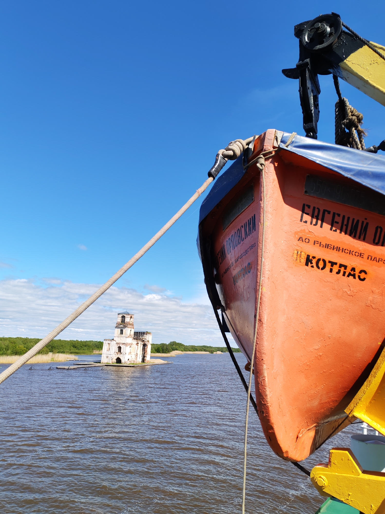 The image depicts an orange lifeboat with the name "Evgeniy Onegin" in Russian on its side, along with "AO Rybinskoye Parokhodstvo" and "Kotlas." The lifeboat is suspended by a black and yellow crane mechanism. A thick rope runs from the top of the lifeboat to the left of the frame.
In the background, there is a body of water, possibly a river or lake. In the middle of the water stands a partially ruined white building with multiple stories and arched windows. The architecture suggests it may have been a church or a religious structure. A green treeline is visible in the far background. The sky is blue with scattered white clouds.
Based on the presence of the ruined church and the style of the boat, this could be a scene near Krokhino, Russia, where a similar flooded church is a known landmark.