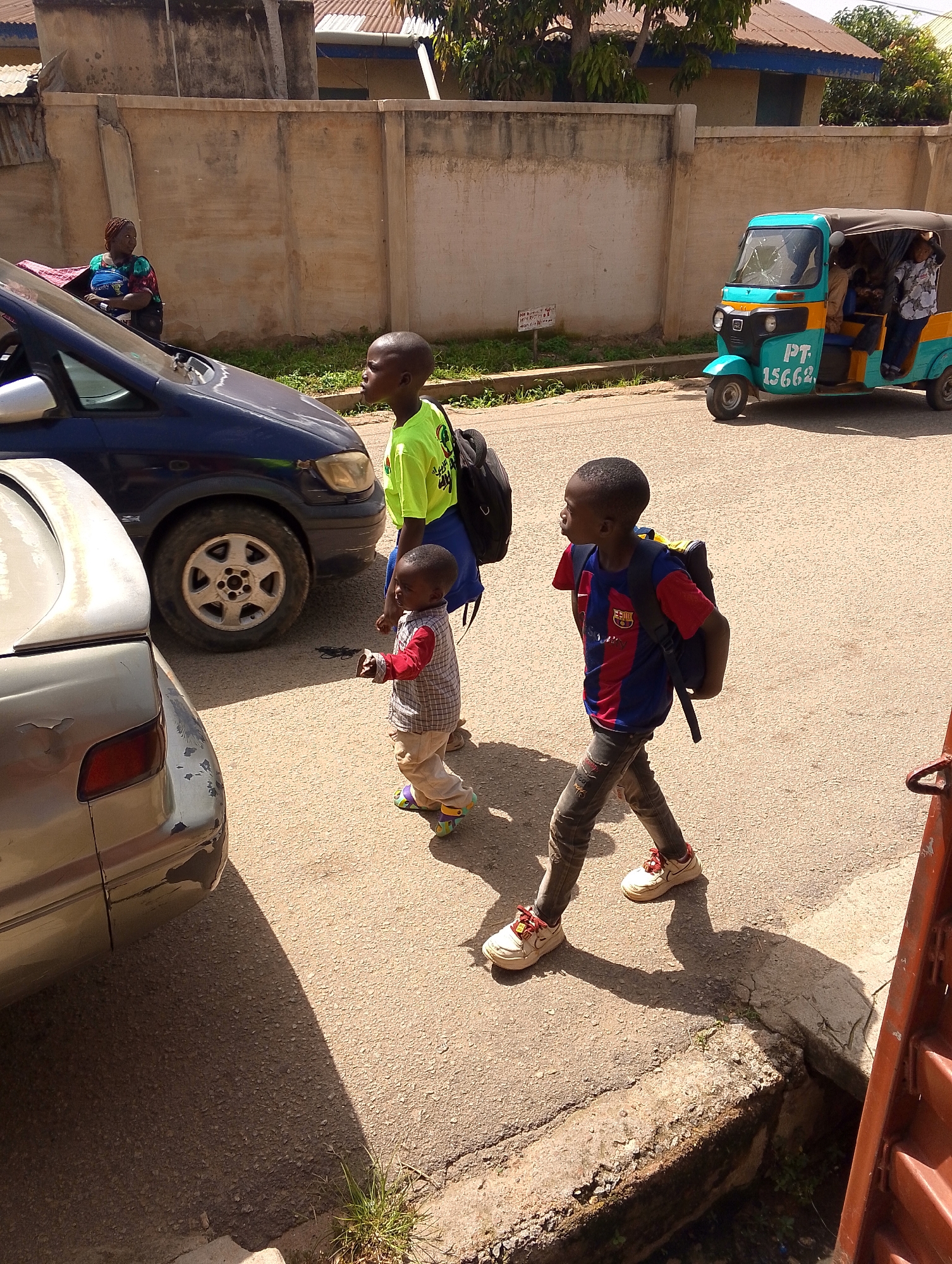 In Jos, Nigeria, on a sunlit day, two young boys walk on a paved street. The boy on the left, wearing a neon green t-shirt and carrying a black backpack, walks with a slight tilt of his head, looking towards the right. Next to him, a younger boy in a plaid shirt and tan pants, with colorful sandals, walks with his arms extended slightly. To the right, another boy in a blue and red striped shirt and distressed jeans, also carrying a backpack, walks forward with his gaze directed ahead. A blue minivan is parked on the left, and a turquoise and yellow auto-rickshaw is in motion on the right. The background shows a textured wall and some vegetation.