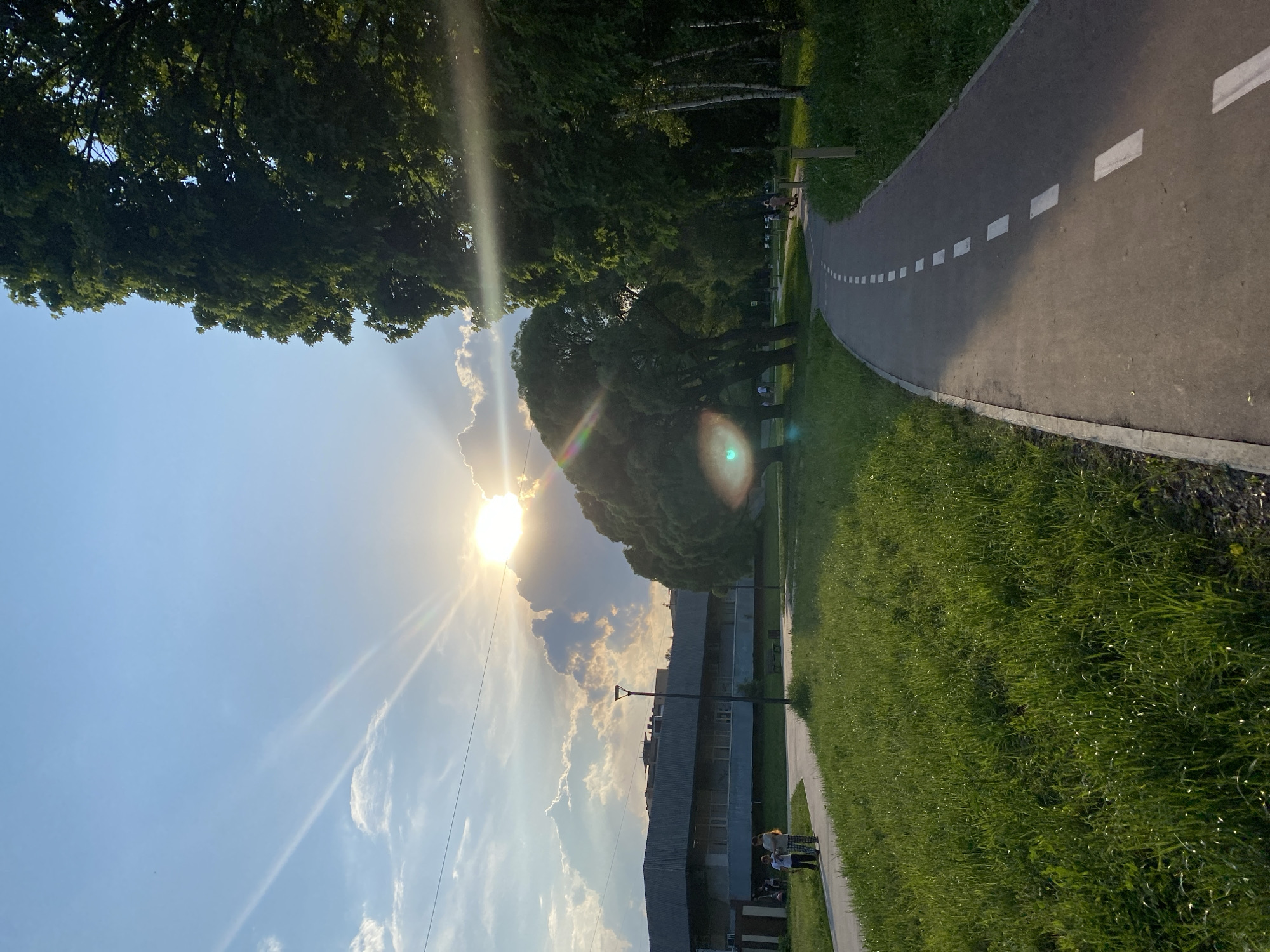 The image shows a park scene in Unknown, Unknown. The sun is obscured by clouds in the sky, casting rays across the sky. On the right side of the image is a paved path with dashed white lines. The path is lined with green grass. In the background, a building can be seen behind trees. People are walking along the path. The tree canopy is visible at the top of the image.