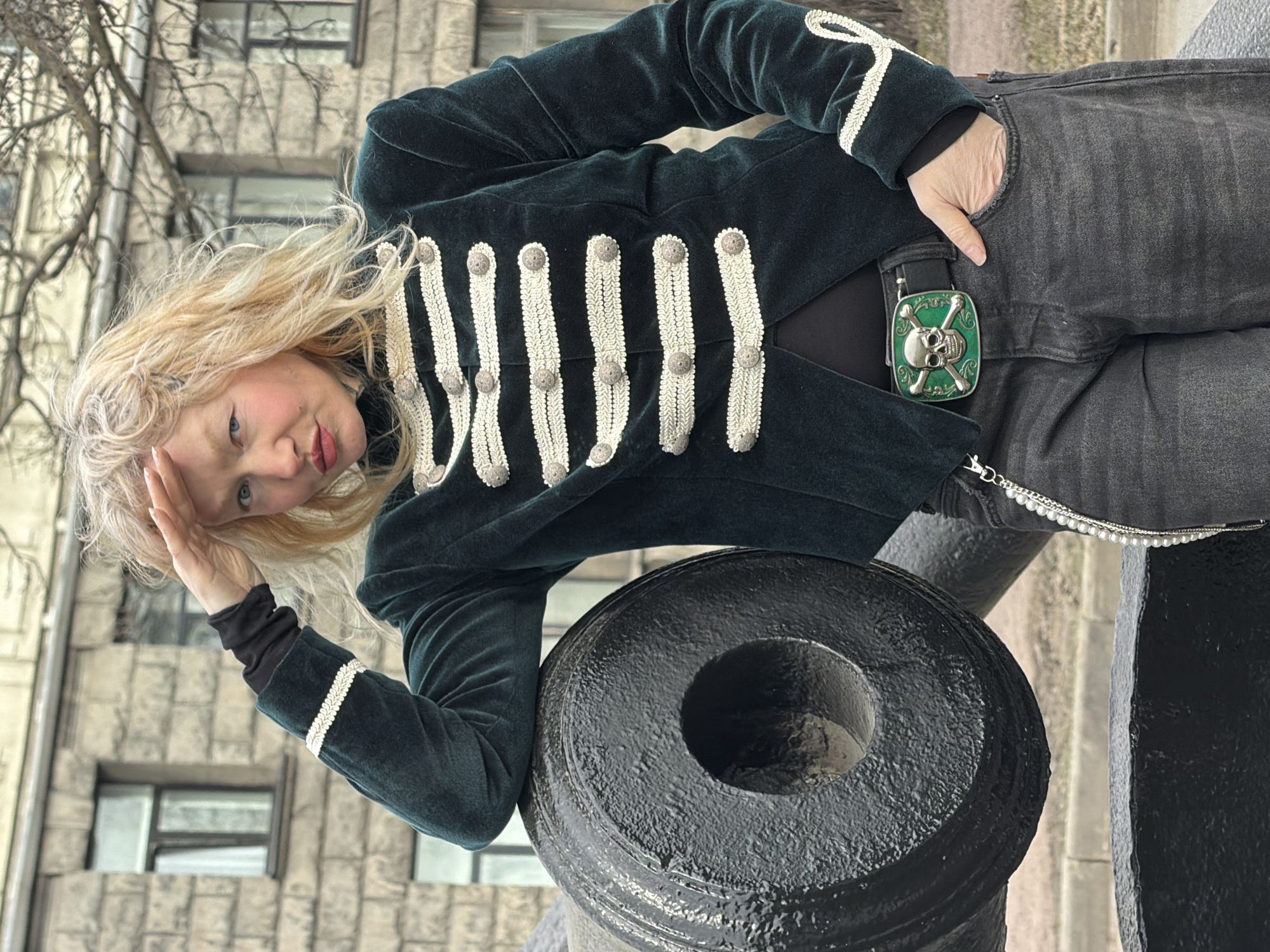A blonde woman is leaning on a black cannon in Saint Petersburg, Russia. She wears a dark green velvet jacket with white trim, black jeans, and a silver skull belt buckle. Her left hand is on her forehead. The cannon is pointed to the right of the frame. A building is visible in the background.
