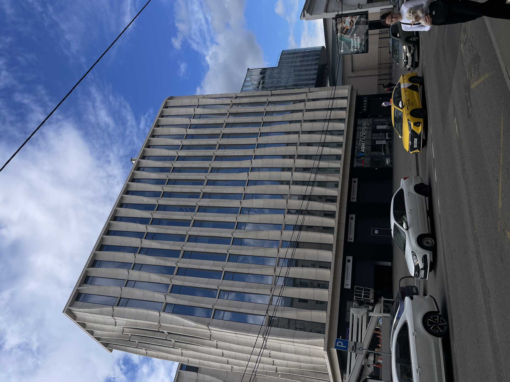 The image shows a street scene in Moscow, Russia, on a partly cloudy day. A modern, multi-story building with a facade of horizontal wave-like concrete elements and numerous windows dominates the left side of the frame. The windows reflect the blue sky and clouds.
Several cars are visible on the street. A prominent yellow taxi with the branding "alo" and Cyrillic text "Яндекс Go" is parked on the right. There are also two white cars, one of which is a hatchback with distinctive wheels, and a darker vehicle between them.
In the background, a woman is holding a small white dog while standing near a sign. The sign, with Cyrillic text, advertises "Отдых в центре города" (Rest in the city center), suggesting a commercial or entertainment area. Another sign on the building reads "ТОРГОВЫЙ ЦЕНТР АФИ ГАЛЕРЕЯ" (Shopping Center AFI Gallery). Numbers 1, 2, and 3 with "Зона разгрузки" (Loading zone) are also visible on the building's lower section, indicating a commercial area with delivery points.
Power lines are strung across the sky and in front of the building. The overall impression is of a typical urban environment with elements of modern architecture and commercial activity. The time of day appears to be daytime, with ample natural light.