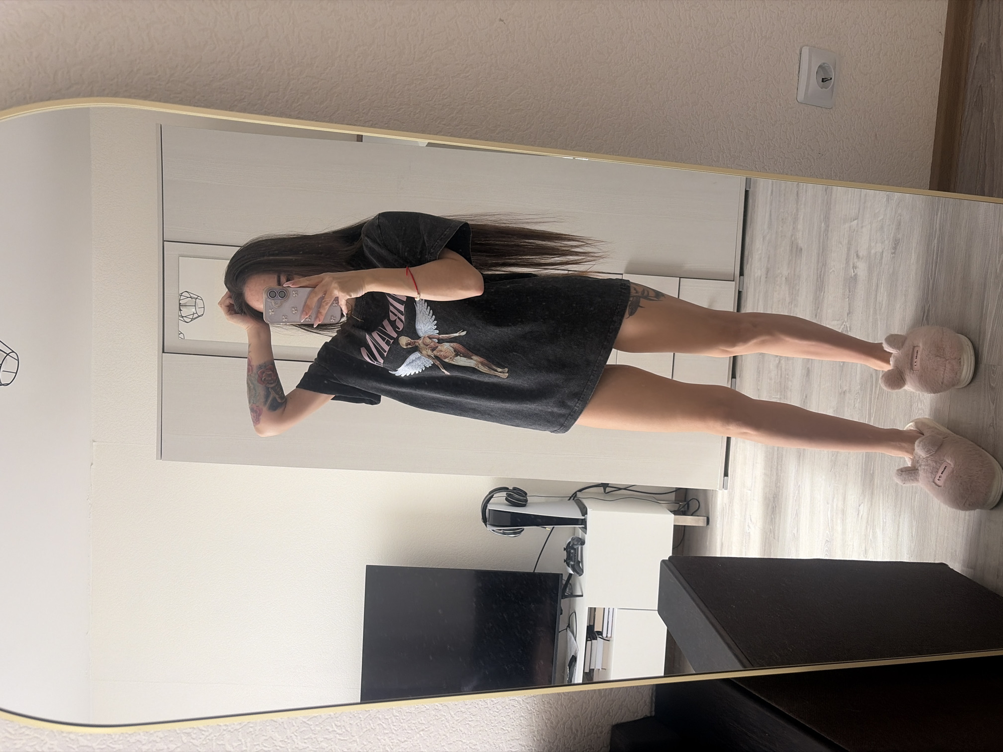 A full-body mirror selfie captures a young person in an indoor setting. The individual, likely female, has long dark hair and is wearing an oversized dark t-shirt featuring a stylized winged anatomical figure and the word "ANGEL" on the front. Visible tattoos adorn their left arm and thigh, and a red string bracelet is on their left wrist. They hold a light purple smartphone with a patterned case to take the photo, largely obscuring their face. Their feet are clad in light pink, fuzzy slippers, one of which has a tag that reads "I <3 dreams."
The setting appears to be a modern room with light-colored, textured walls and light wooden or laminate flooring. Reflected in the mirror behind the person are various objects: a white wall-mounted electrical socket, a white entertainment unit or set of shelves, a black flat-screen television, a white PlayStation 5 console with headphones draped over it, and a game controller. A framed picture with a black abstract drawing hangs on the wall. A dark, rectangular object, possibly a speaker or ottoman, partially blocks the lower right corner of the mirror's reflection. The mirror itself has a light, possibly gold-toned, frame. The overall impression is a casual, domestic scene.
