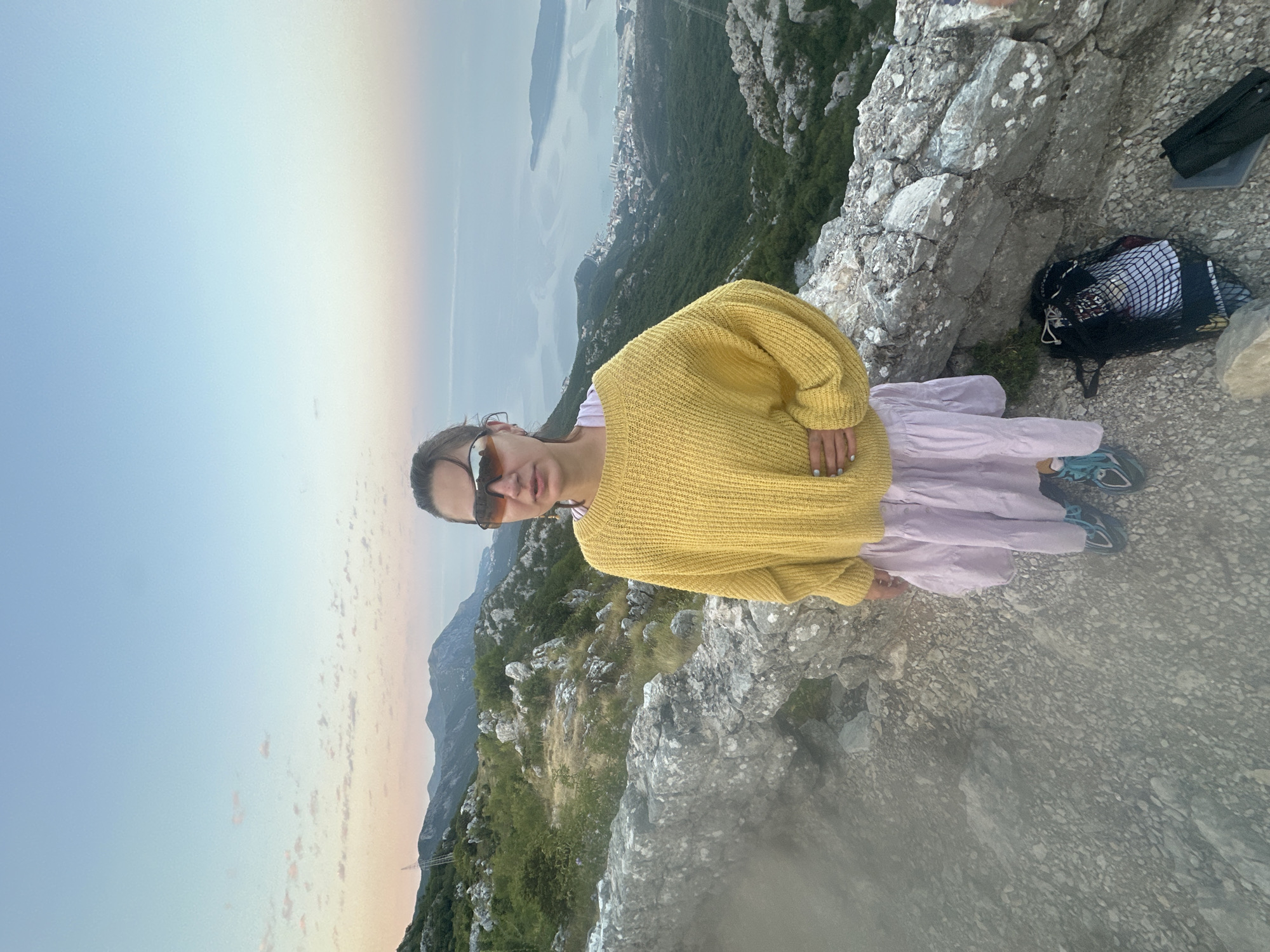 A woman stands on a rocky incline overlooking a coastal town at dusk in Brajici, Montenegro. She is wearing a yellow knitted sweater, a pale pink skirt, sunglasses, and athletic shoes. Her right hand is on her hip. A black mesh bag and a black case are on the ground beside her. The landscape features rolling hills covered in vegetation, a body of water with scattered islands, and a distant, pale sky with wisps of clouds. The foreground is dominated by rough, gray stones and gravel.