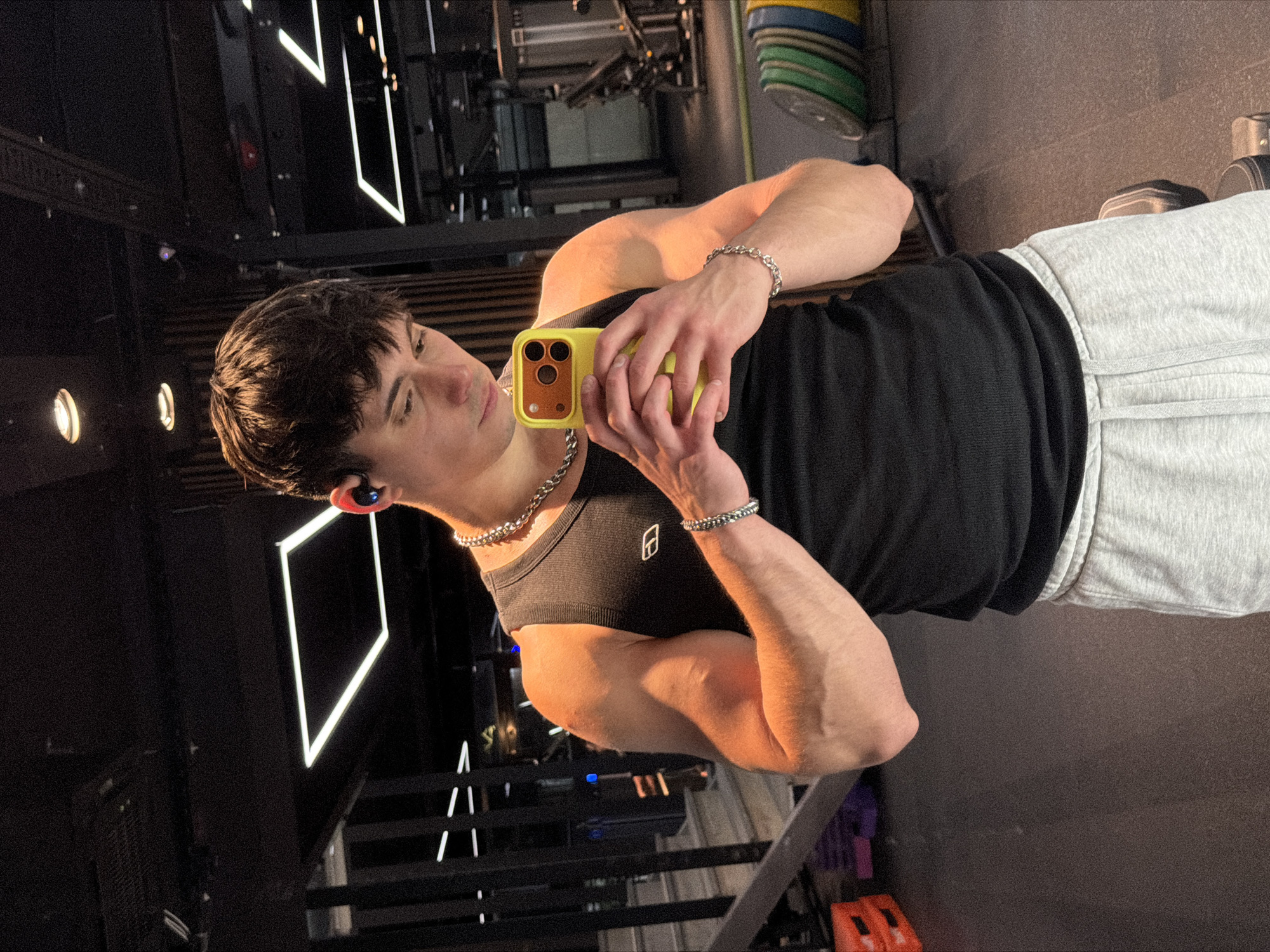 A young man is taking a selfie in a gym setting. He is wearing a black tank top and grey sweatpants, with a silver chain necklace and bracelet. He holds a yellow phone in his hands, using it to capture the image. An earpiece is visible in his ear. The gym environment features various fitness machines and weights. Geometric light fixtures are present on the ceiling. The scene appears to be indoors, in a commercial gym setting. There is no location information in the picture itself so it cannot be determined if it is in the city of Sofia, Bulgaria.