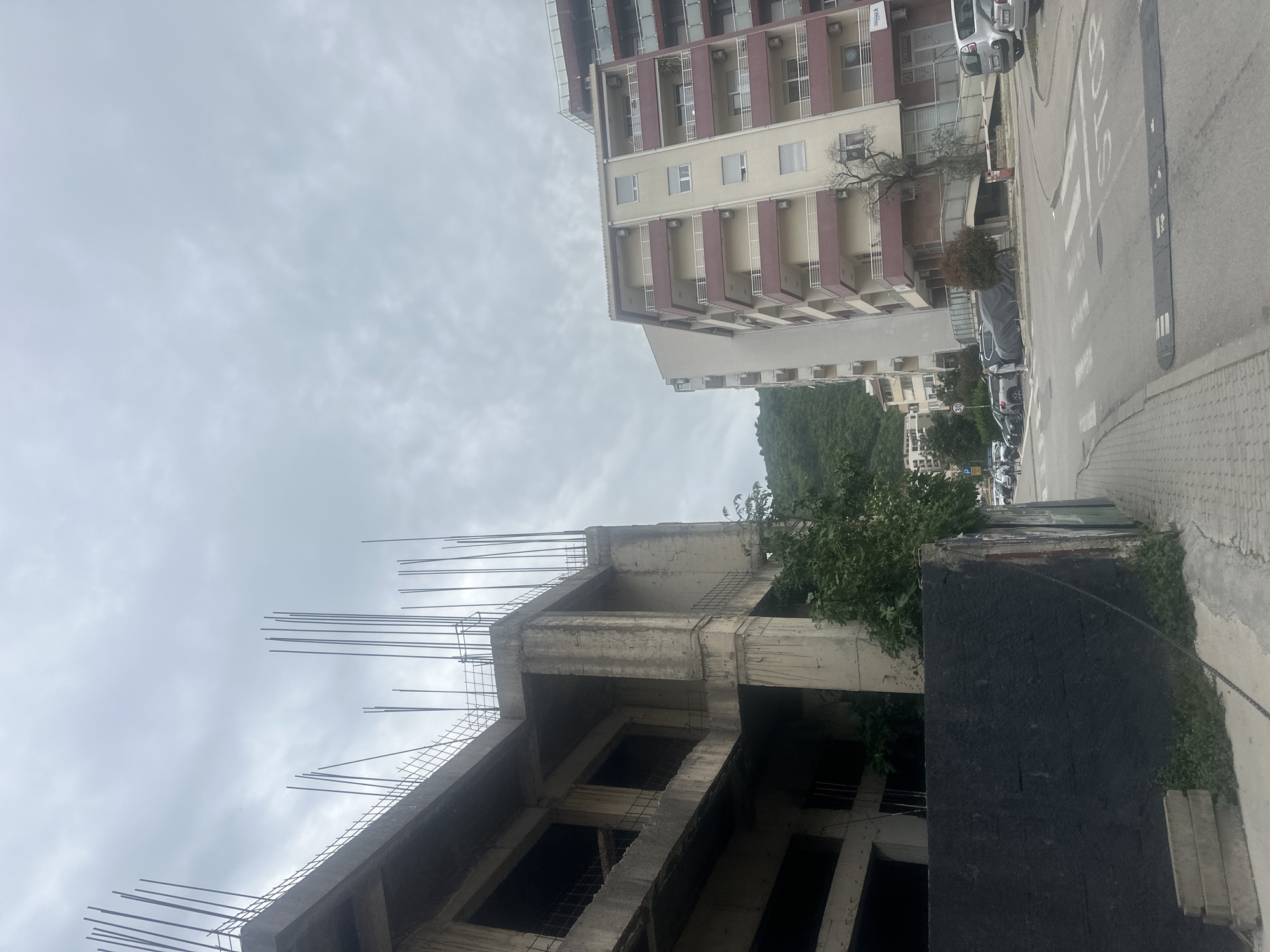 The image captures a street scene in Budva, Montenegro, featuring an unfinished concrete building with exposed rebar extending skyward. Adjacent to it stands a completed apartment building with balconies and windows. The street is paved, with cars parked along the side, and a pedestrian crossing is visible. The sky is overcast. The scene shows a blend of ongoing construction and completed urban infrastructure.