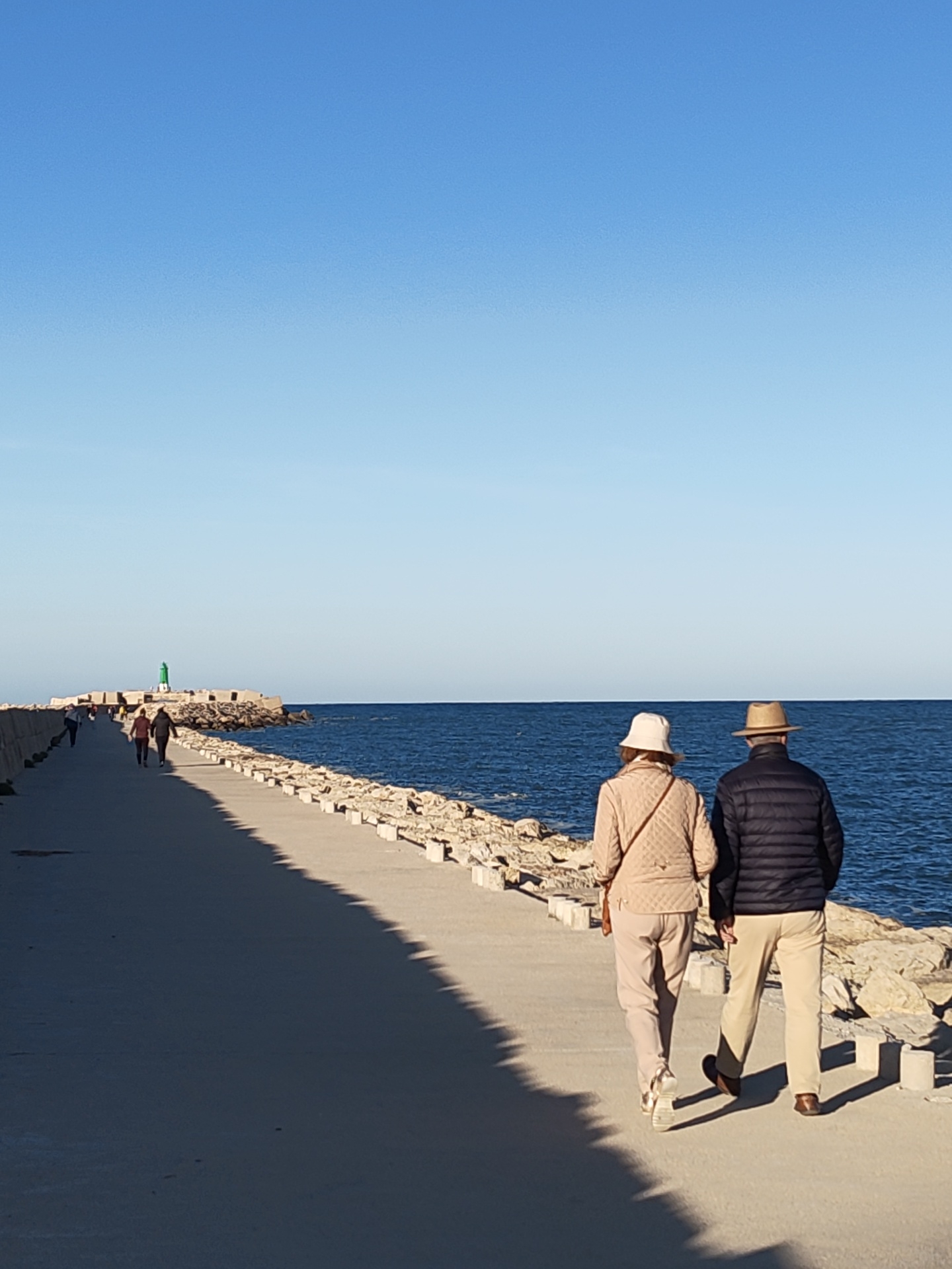 The image captures a sunny scene on the northern breakwater (Dique de Poniente) of the Port of Dénia, Spain. A wide concrete pier extends into the deep blue sea, leading towards a green lighthouse visible at its distant end. The pier's right edge is lined with a rock embankment and concrete blocks. Sunlight from an angle to the left casts long, distinct shadows across the pier.
In the foreground, an older couple walks away from the viewer. The woman wears a light-colored quilted jacket, light-colored trousers, a white bucket hat, and light footwear; a small cross-body bag is visible. The man wears a dark puffer jacket, light-colored trousers, a straw fedora, and dark loafers. Several other individuals are visible walking further down the pier towards the lighthouse. The sky above is clear and blue.