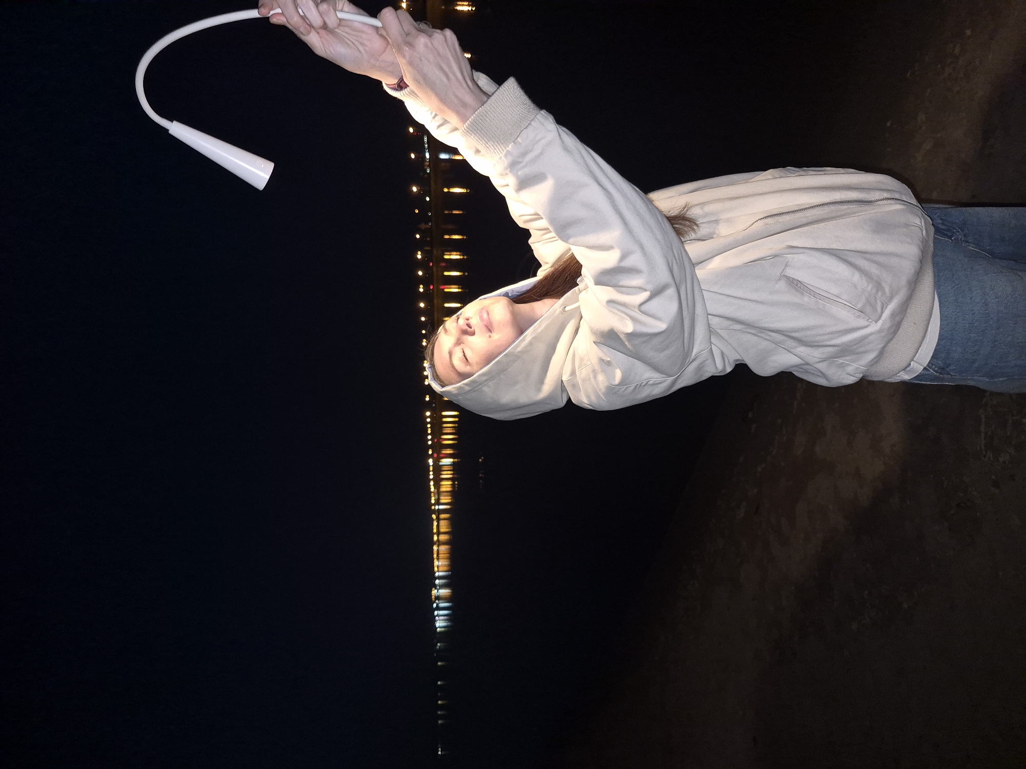 A woman in a hooded jacket stands against a dark background, illuminated by city lights in the distance, holding a white flexible lamp. She appears to be outdoors near a body of water or a dark, paved surface. The city is Unknown, Unknown.
