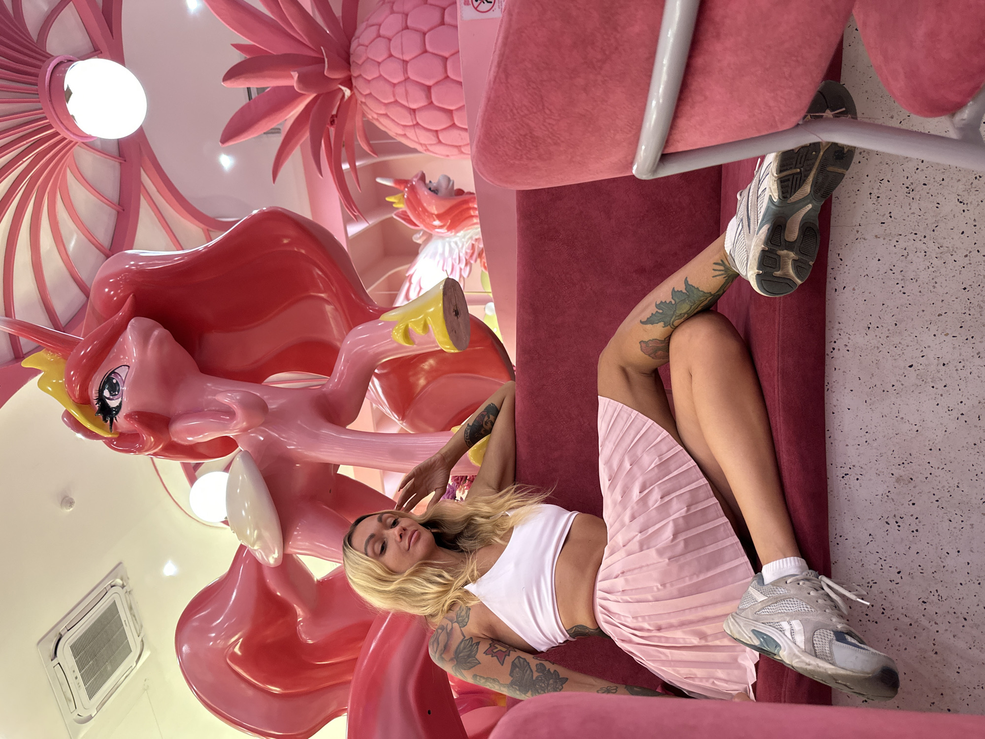 A blonde woman with tattoos on her arms and legs is sitting on a pink sofa in a cafe. She is wearing a white crop top and a pink pleated skirt. The cafe is decorated with pink cartoon figures, including a large pink unicorn and a pink pineapple. The woman is looking at the camera with a slight smile.