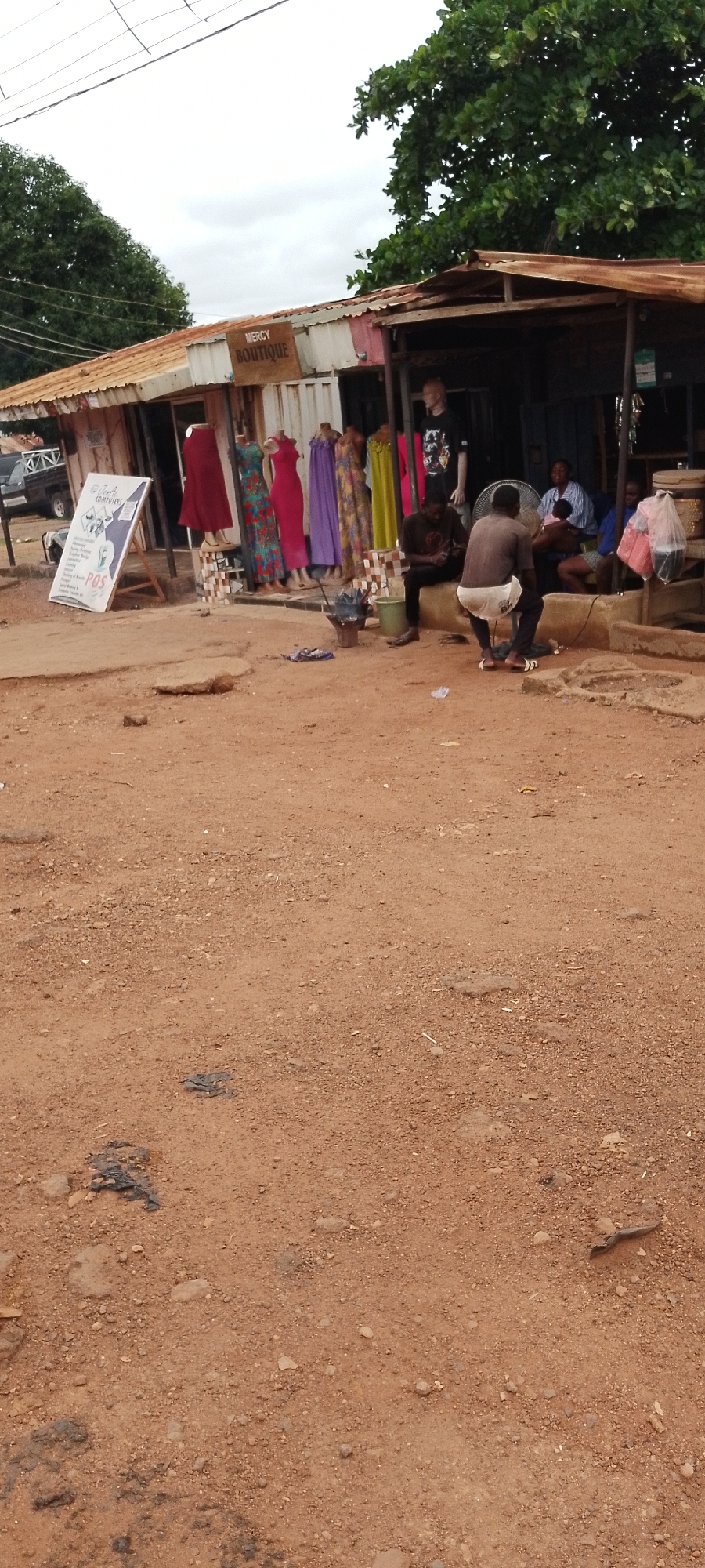 This image shows a street scene in Gboko, Nigeria, likely a market area. In the foreground, the ground is covered in reddish-brown dirt with scattered stones and debris. A small black object, possibly trash, is visible on the ground.
Dominating the middle ground is a stall with a corrugated metal roof. The stall displays several colorful dresses on mannequins, with various fabrics like patterned, red, purple, and yellow visible. Above the stall, a sign reads "MERCY BOUTIQUE". To the left of the stall, a white A-frame sign advertises "Thistle Computers" and "POS".
In the background, under the shelter of the stall, several individuals are present. One person is sitting and looking down at a mobile device. Another person, a woman, is holding a baby. A man is seated next to her. In front of them, a black electric fan is positioned. To the right of the fan, a person wearing a brown t-shirt and white shorts is squatting and appears to be looking down. They are wearing sandals.
Further to the right, more merchandise is visible, including items wrapped in pink plastic bags. A dark-colored vehicle, possibly a pickup truck, is parked to the far left, partially obscured. The environment is characterized by a rustic, open-air setting with a few trees visible in the upper right portion of the image. The sky is overcast.