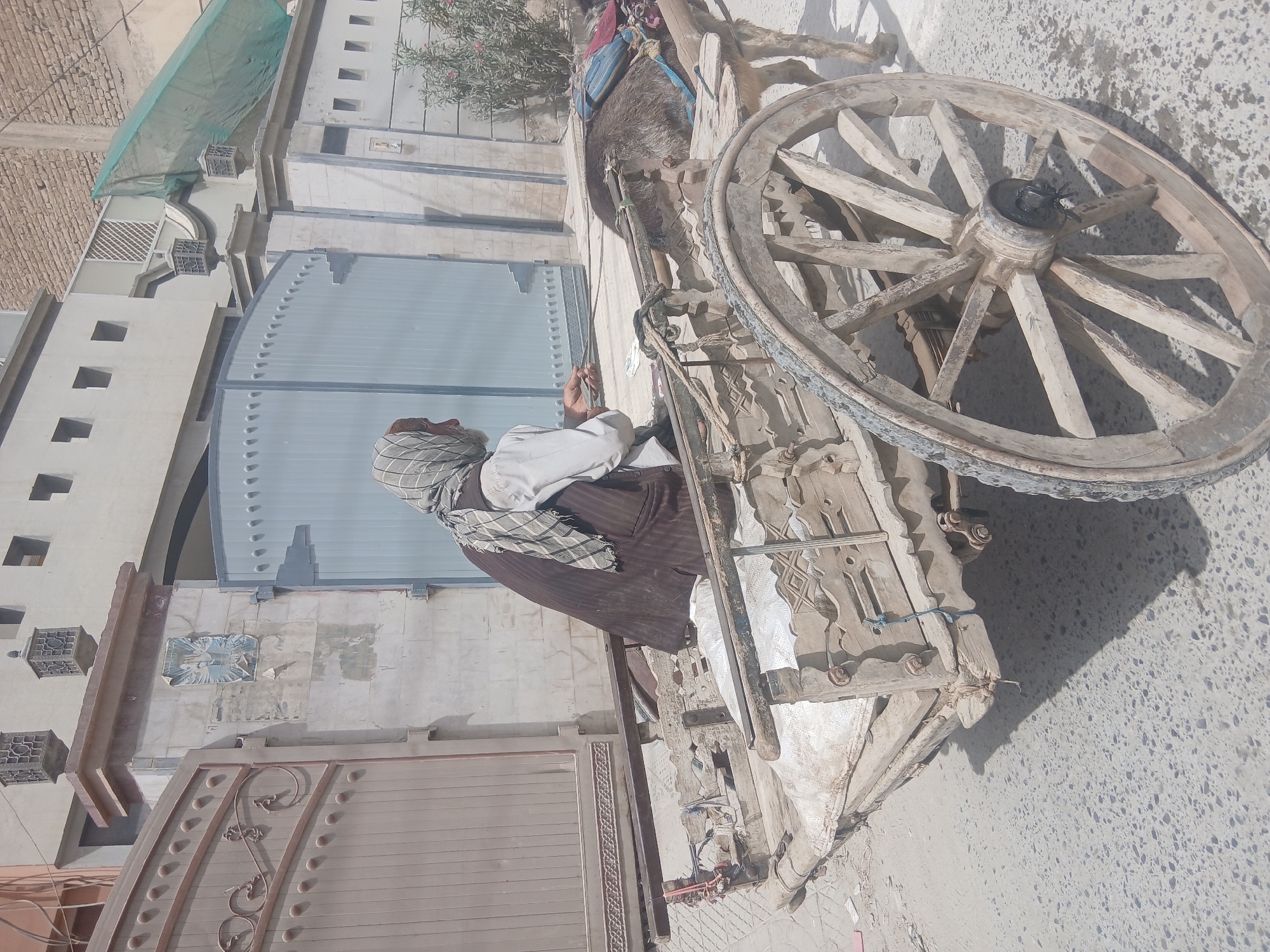 A man is on a donkey cart in Quetta City Tehsil, Pakistan. The cart is old and wooden, with large wooden wheels. The man is wearing traditional clothing and a head covering. The background includes buildings with a blue gate. The overall environment appears to be a street or alleyway in a city.