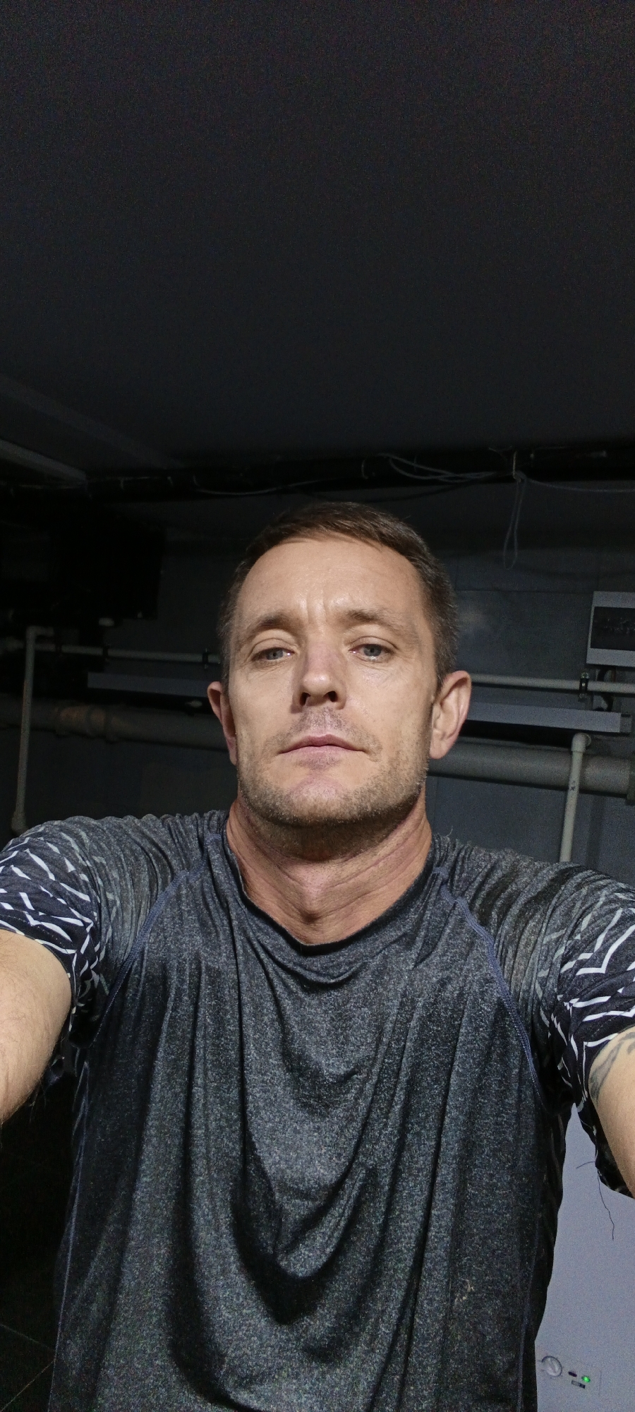 A man, appearing to be in his late 30s or early 40s, is shown in a close-up, head-and-shoulders portrait. He has short, brown hair, light blue eyes, and stubble on his chin and jawline. He is wearing a dark grey t-shirt with a black and white patterned design on the shoulders and sleeves. The background is dark and appears to be indoors, with visible pipes and wires suggesting an industrial or utility area. There is no indication of the specific location within Tashkent, Uzbekistan. The man is looking directly at the camera with a neutral expression.