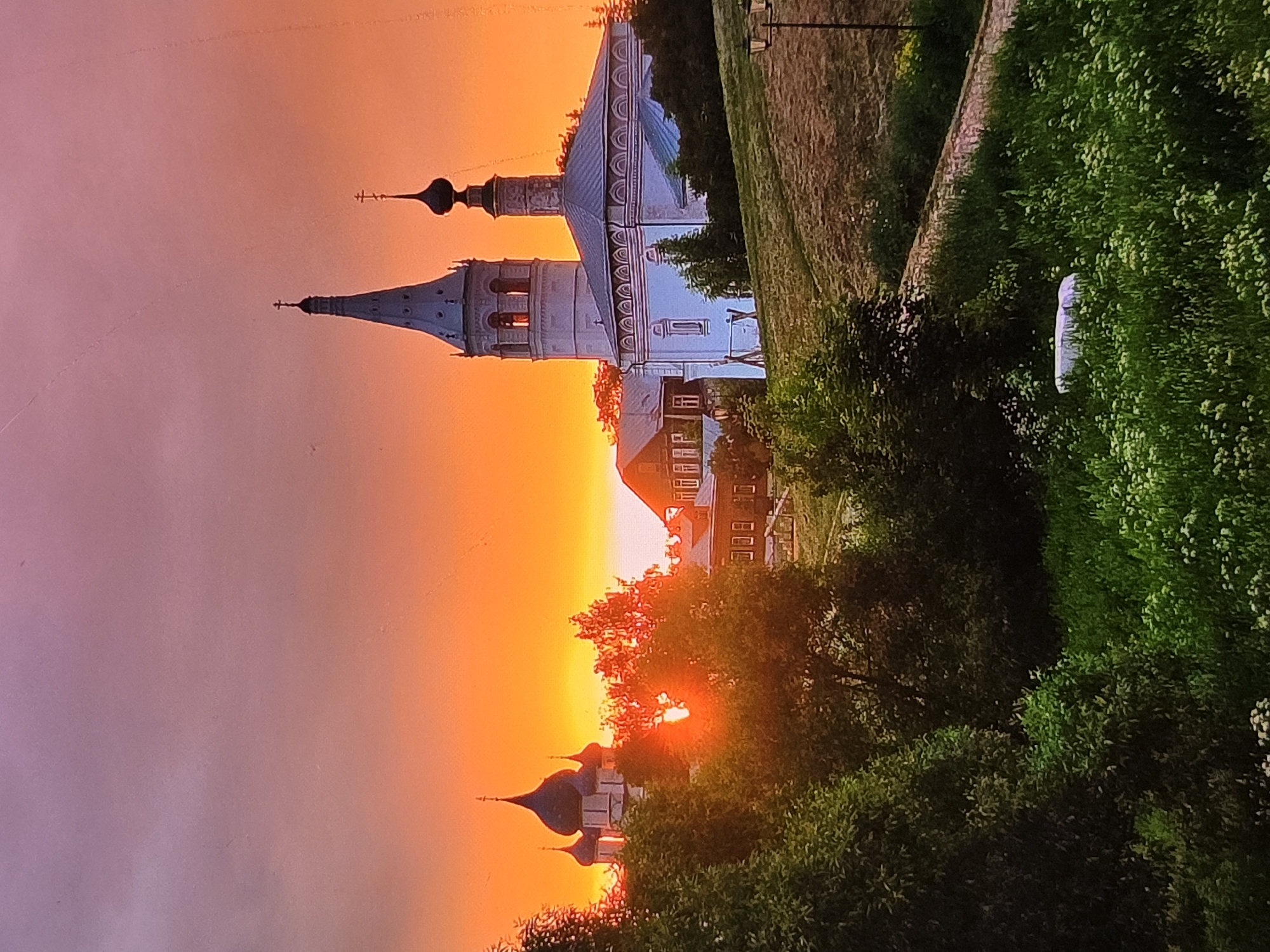 The photograph captures a sunset over the city of Vladimir, Russia. In the foreground, lush green grass and trees are illuminated by the warm, orange glow of the setting sun. The sun's rays are visible, peeking through the foliage of a tree, casting a bright light.
In the midground, several domed churches and traditional Russian wooden houses are nestled amongst the trees. The church on the right, with its distinctive white walls and blue-grey roof, stands prominently. To its left, another smaller church with dark blue domes is partially obscured by trees. The sky above is a gradient of vibrant orange and soft pink, indicative of a dramatic sunset.
Further in the background, the rolling hills suggest a slightly elevated viewpoint. The scene is tranquil, with no people or animals visible, presenting a serene, almost idyllic, depiction of a Russian landscape at dusk. The overall impression is one of natural beauty and architectural heritage.