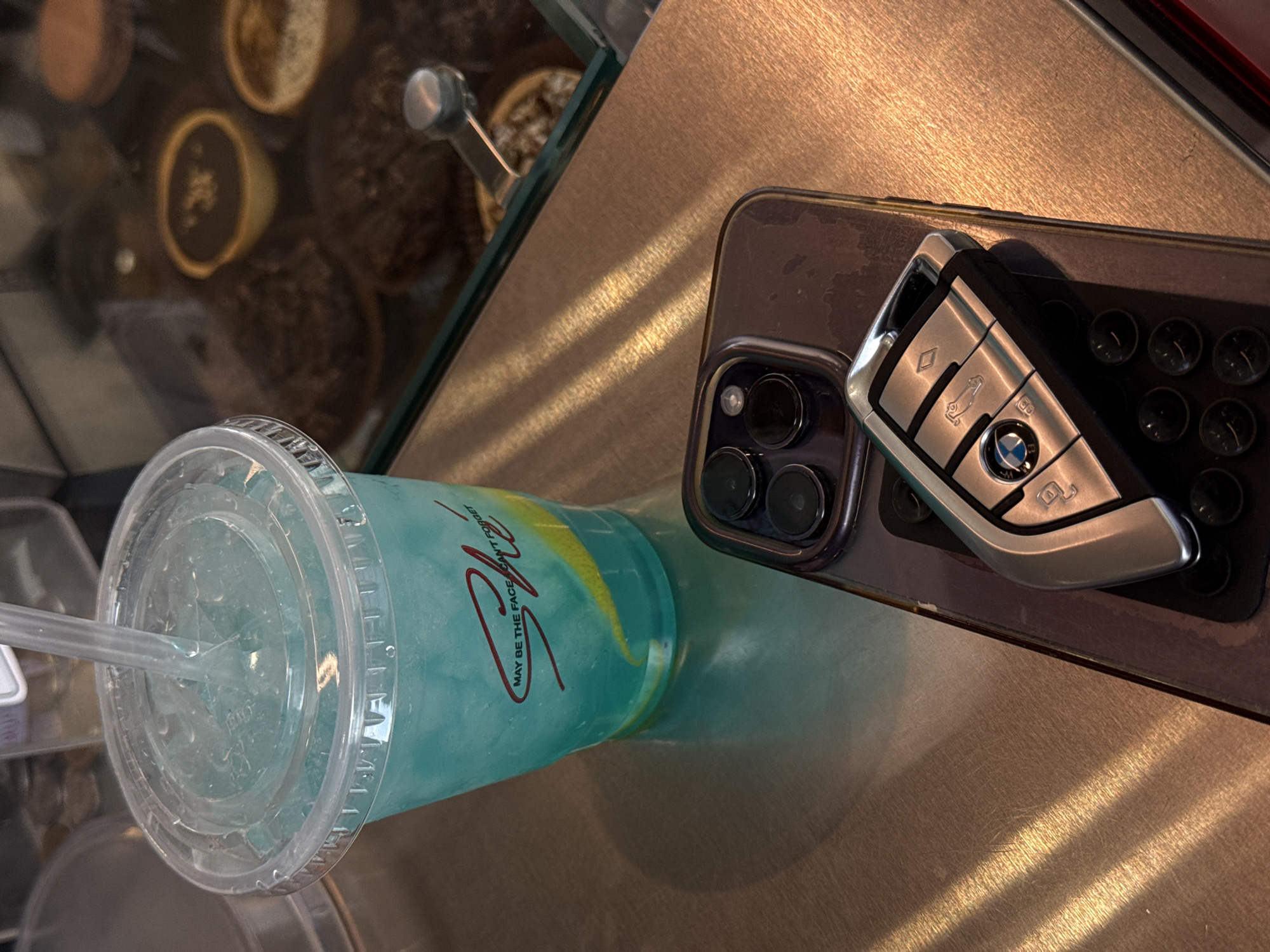 The image shows a blue drink with a clear lid and straw next to a phone and BMW car key. The drink has the logo "May Be The Face I CAN'T FORGET" on it. The objects are on a metal-look surface. The location is Yerevan, Armenia.