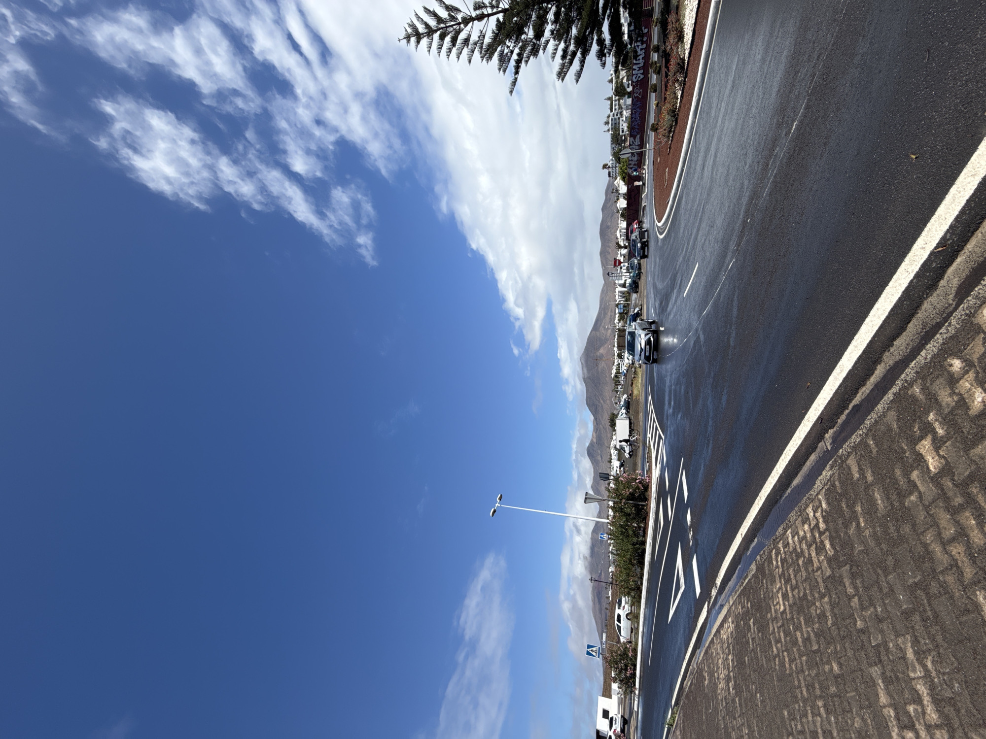 The photograph depicts a road in Yaiza, Spain, characterized by a sharp curve. The road surface appears wet, possibly due to recent rain, and has white lane markings. A variety of cars are visible, some parked along the roadside. In the background, there is a mountain range. The sky is a gradient of blue, ranging from a clear, deep blue to areas with scattered white clouds. A light post is visible along the roadside. A sidewalk made of interlocking pavers lines the edge of the road.