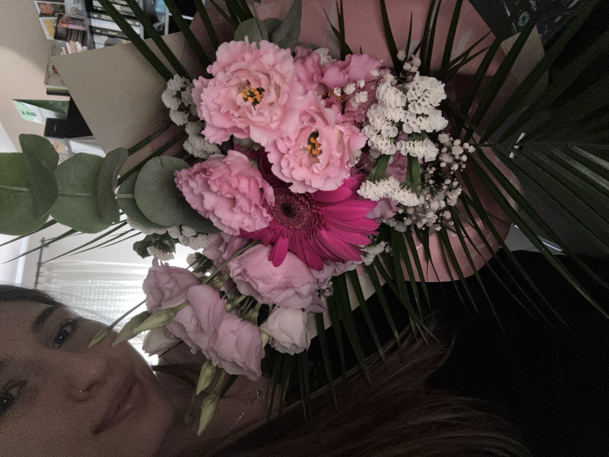 The image shows a close-up of a person holding a bouquet of pink and white flowers. The person, a young woman, is partially visible in the upper left corner of the frame, with her face turned towards the camera. She has dark hair and is wearing a black top. The bouquet is comprised of various pink roses, a vibrant pink gerbera daisy, and smaller white flowers, possibly baby's breath. Green foliage, including palm-like leaves and eucalyptus branches, is also incorporated into the arrangement. The background is blurred but appears to be an indoor setting with bookshelves and curtains visible. There is no indication of the city of Veliko Tarnovo, Bulgaria, or any specific location.