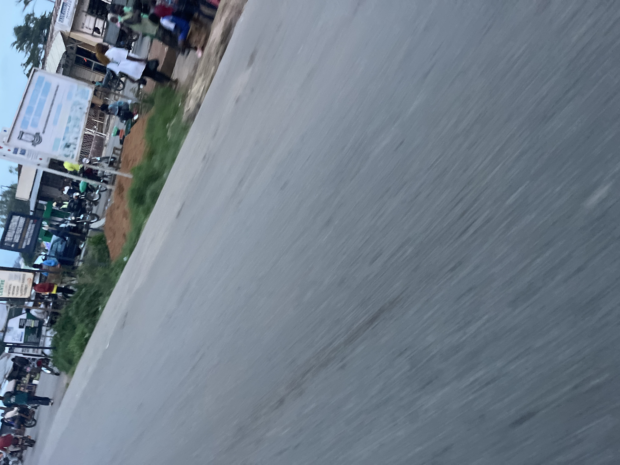 The image captures a street scene in Gura Top, Nigeria, with a focus on the movement blur of a paved road in the foreground, indicating motion. The background depicts a busy roadside environment with several people, motorcycles, and market stalls. On the left side of the frame, individuals are seen near parked motorcycles. Further into the scene, there are structures that appear to be shops or market stalls, with signs visible, including one with the word "CENTRE". People are engaged in various activities, some standing, some walking, and others seated. One person in a white shirt is walking with a dark bag. A blue car is partially visible in the background. The overall impression is of a typical urban street in Nigeria, with a lively atmosphere and everyday activities taking place.