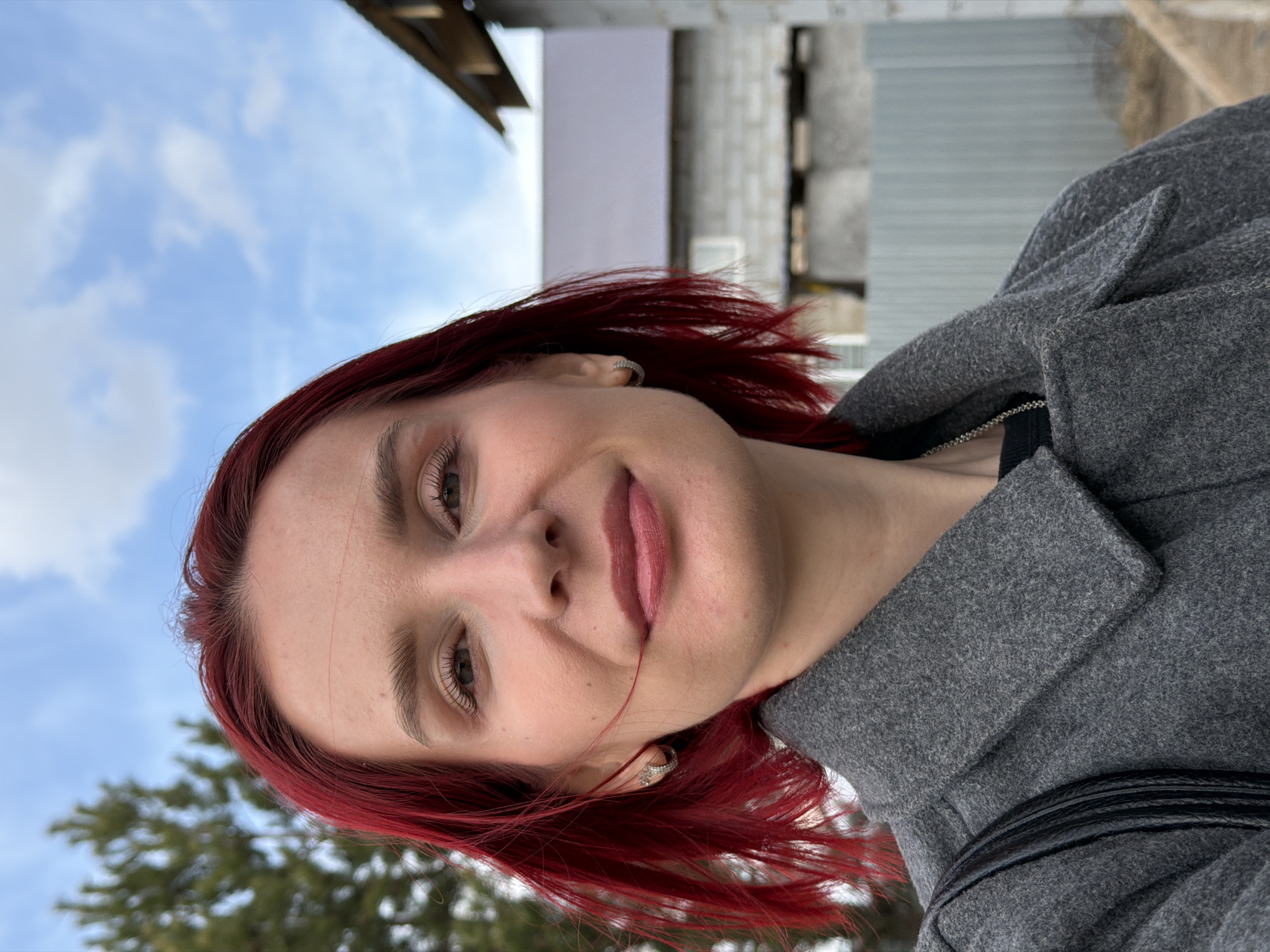 The image displays a head-and-shoulders portrait of a young woman taken outdoors in Chaikovsky, Russia. The photograph is oriented 90 degrees counter-clockwise.
The subject is a young woman with distinctive, bright red hair, worn loose and slightly tousled. She is looking directly at the camera with a soft, gentle smile, her lips slightly parted. She has subtle makeup, including visible mascara and eyeliner, and a light lip color. Small silver stud or hoop earrings adorn her ears.
She is wearing a textured, medium-gray coat with a structured collar, suggesting cool weather. A black garment is visible beneath the coat, around her neck area.
The background consists of a clear blue sky with some scattered white clouds. To the lower left, the blurred branches of a coniferous tree are visible. On the right side of the frame, there is a building or structure made of gray concrete blocks and a lighter-colored section, possibly indicating a residential or industrial setting. The overall impression is of a daytime outdoor scene. No visible text is present in the image.