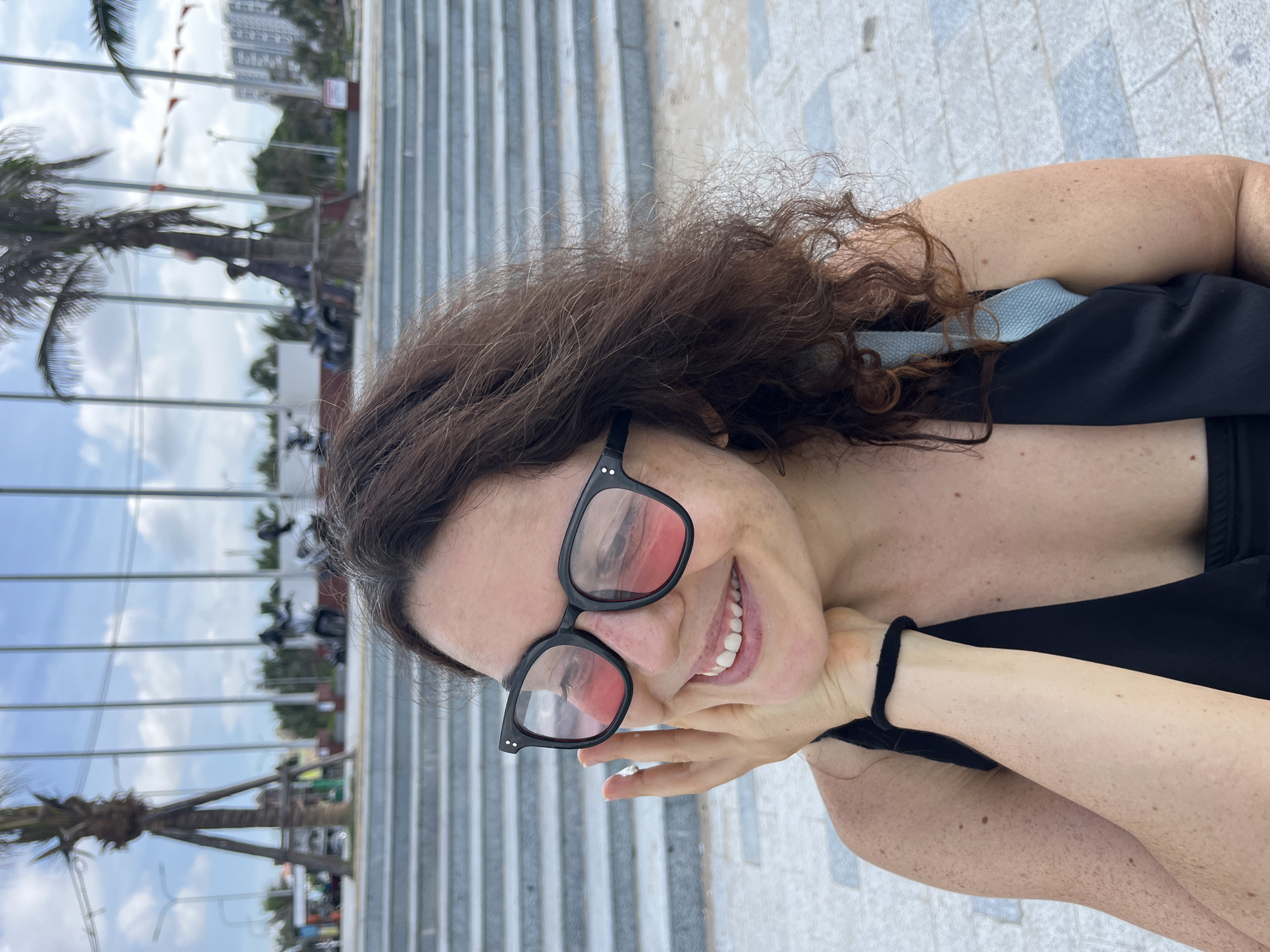 A medium shot captures a woman with brown curly hair and freckled skin, wearing black-framed sunglasses with pink lenses and a black top. She is outdoors, touching her face with her right hand, and smiling. The background features a paved walkway, a set of tiered concrete stairs, palm trees, and distant buildings and vehicles under a cloudy sky. This scene is likely located in Phan Thiết, Vietnam, near a public area with outdoor seating and pathways.