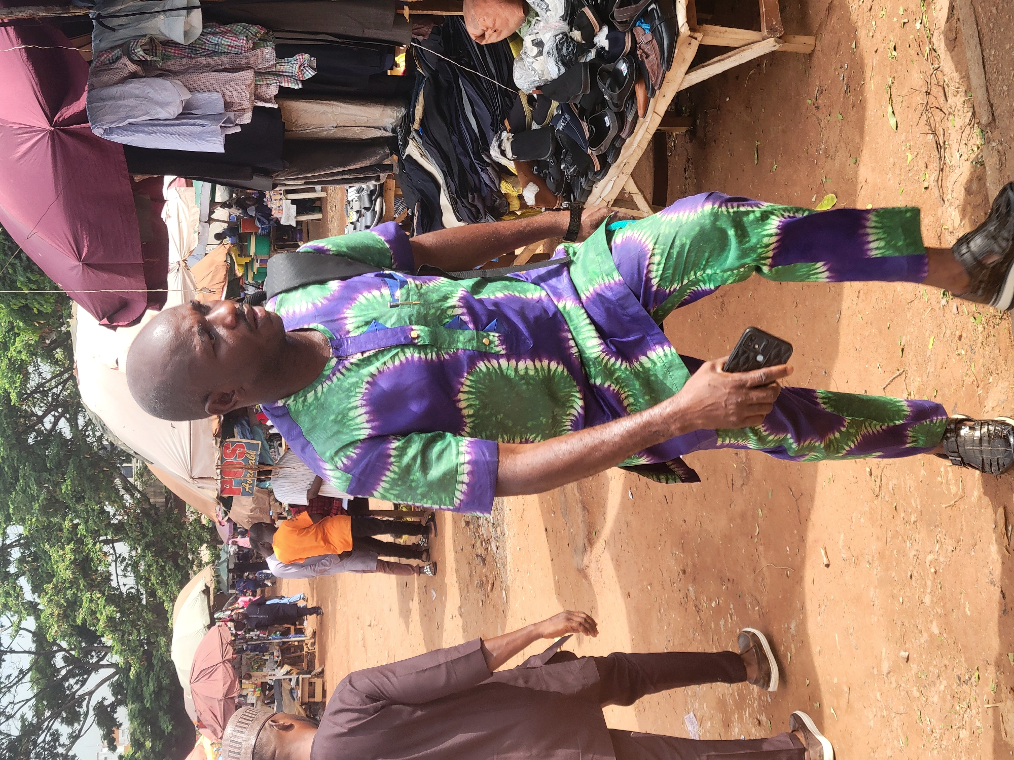 The scene is an outdoor marketplace in Abuja, Nigeria. The ground is reddish-brown dirt, possibly clay. In the foreground, a man with a shaved head is sitting on a makeshift wooden structure. He is wearing a green and purple patterned shirt and pants set and black shoes. He is holding a smartphone in his left hand. Behind him, on the wooden structure, are piles of clothing and shoes for sale. Various people are walking by in the background. Parasols and canopies cover the market stalls. A man in a brown outfit is walking past in the lower part of the frame.