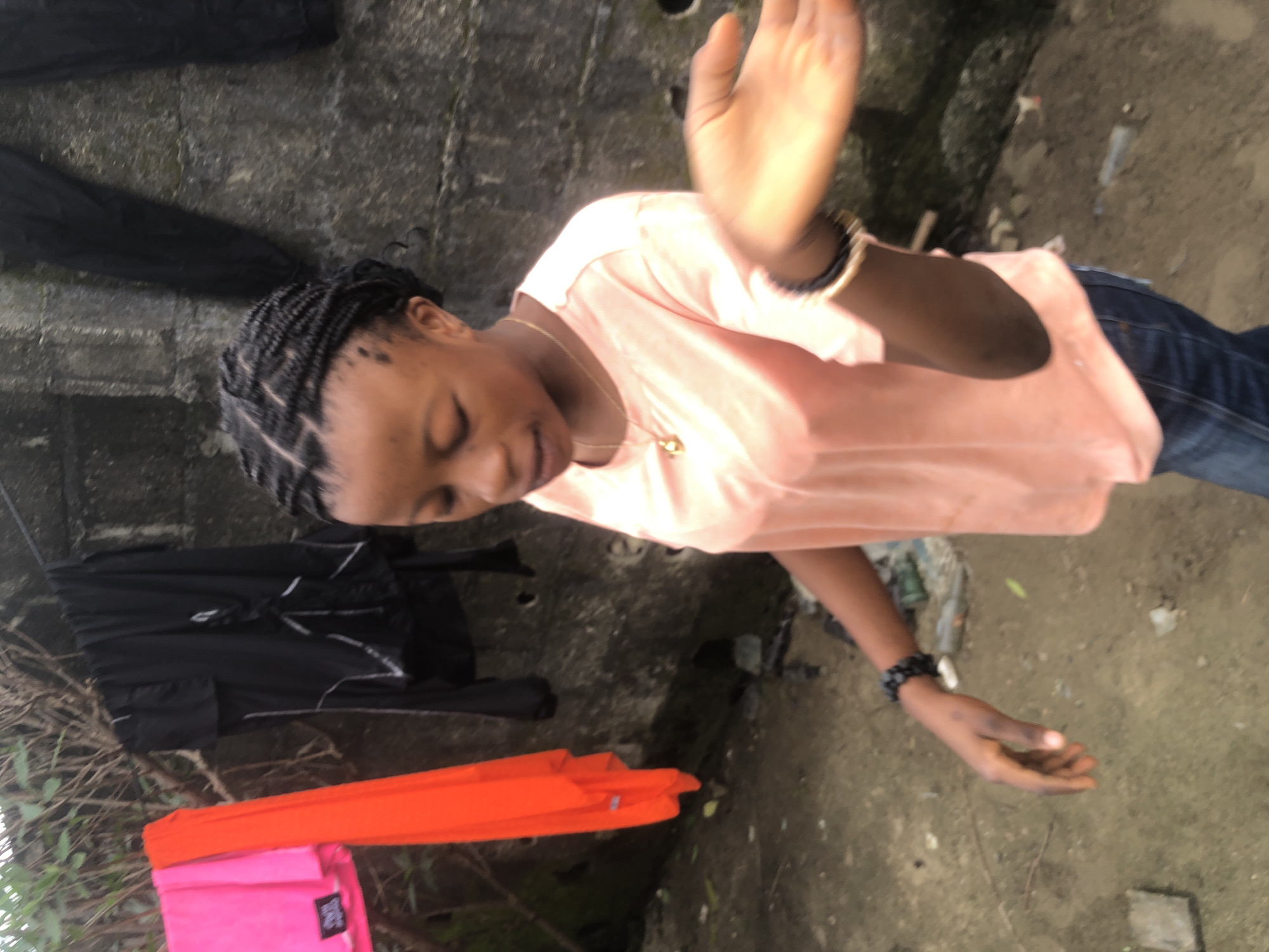 A young Black female with braided hair and wearing a peach-colored t-shirt and blue jeans stands outdoors in what appears to be a residential area in Ado, Nigeria. She is looking down and to her right, with her left arm bent and her hand extended slightly, palm open. She wears a gold pendant necklace and two bracelets on her left wrist. In the background, laundry hangs on a line, including a bright pink and an orange garment, as well as black clothing. A textured concrete wall is visible behind the laundry. The ground is dirt and littered with debris, including plastic bottles and what appear to be pieces of wood or concrete. The lighting suggests an overcast day.