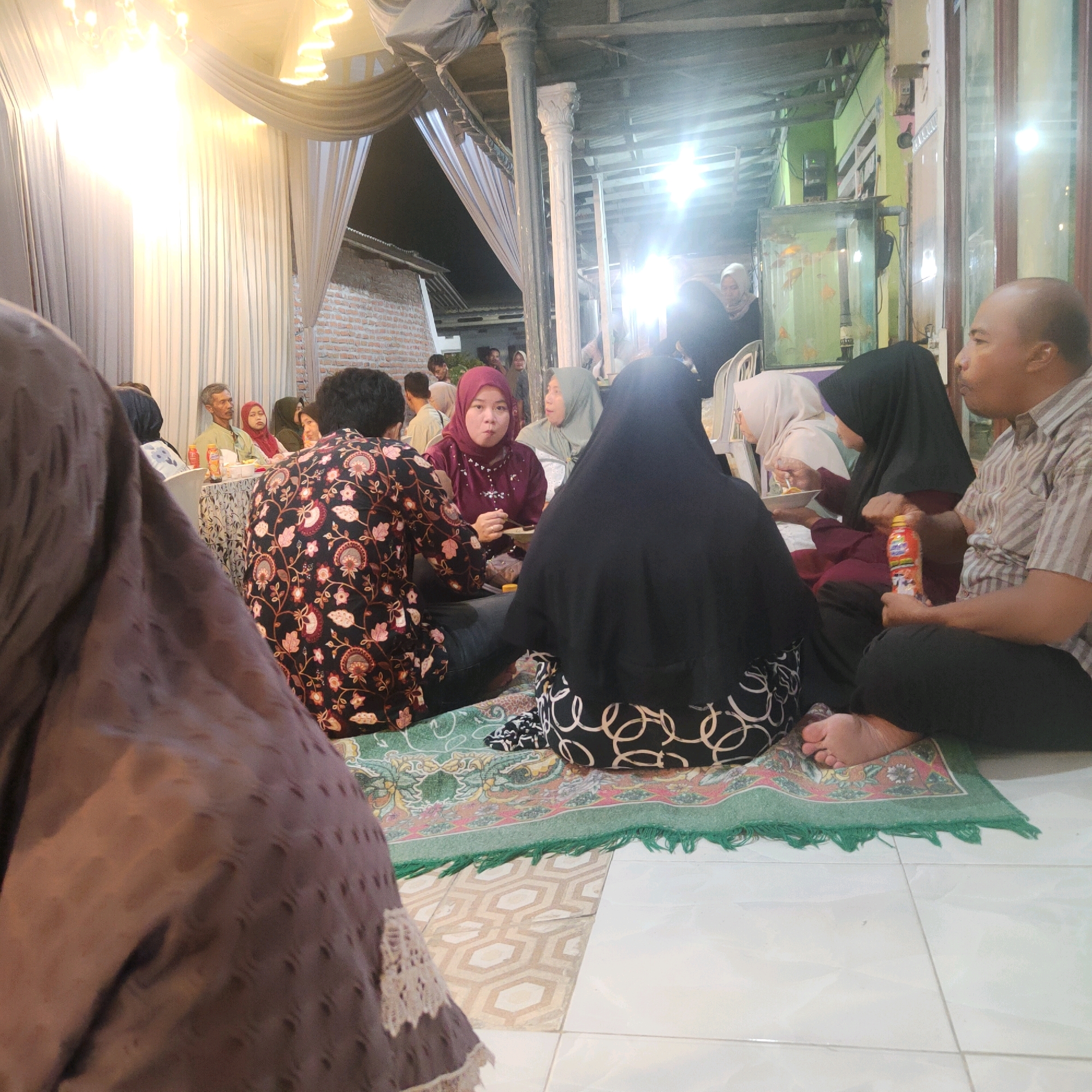 The image depicts an indoor gathering, possibly a meal, in Kolursari, Indonesia. Several people are seated on the floor on rugs, with some at a low table laden with food and drinks. The room appears to be decorated with draped fabric and is illuminated by overhead lights. In the background, a brick wall and a fish tank with orange fish are visible, suggesting a residential or community space. The people are dressed in traditional attire, including headscarves. A man in the foreground is holding a beverage bottle, while others are engaged in eating or conversation. The overall atmosphere seems casual and communal.