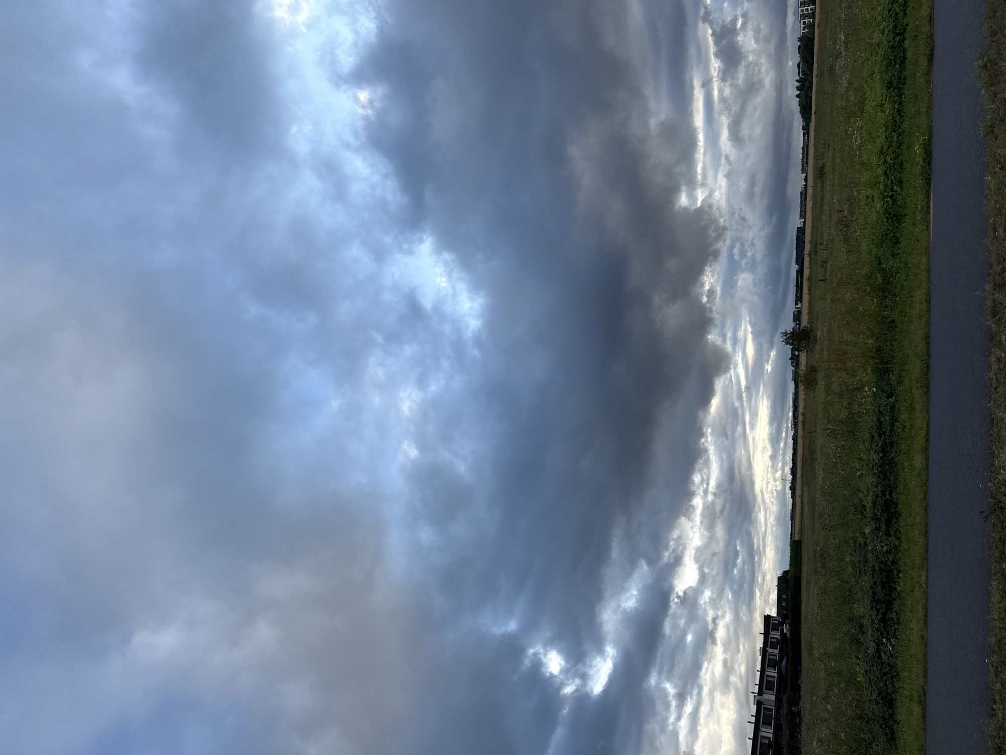 The image depicts a view of Peetri alevik, Estonia, featuring a dramatic sky filled with heavy, dark clouds, interspersed with patches of lighter blue. The foreground shows a paved pathway with a strip of green grass bordering a wide field of tall, dry grass and wildflowers. In the mid-ground, a line of modern, multi-story residential buildings with dark facades and light-colored windows is visible along the horizon, stretching across the landscape. Further in the distance, more buildings and trees are scattered, with faint hints of other structures and a radio mast to the right. The overall scene suggests an overcast day, possibly before or after a rain shower, with a suburban or semi-rural setting. There are no people or animals visible in the frame.