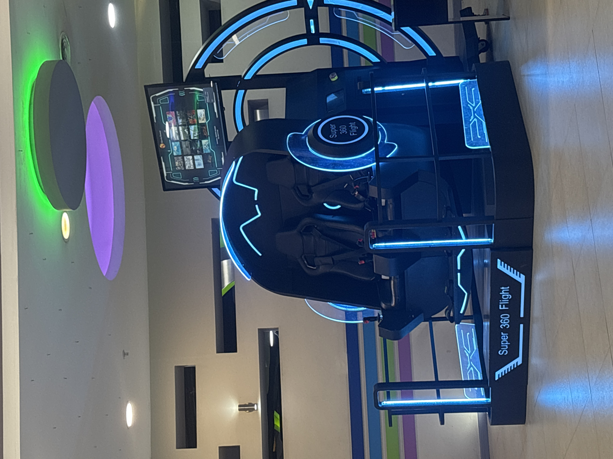 This image captures a vibrant virtual reality (VR) or simulation ride located indoors, likely within an entertainment center or mall in Yerevan, Armenia. The central subject is a dark-colored, two-seater simulation machine heavily accentuated with striking blue neon or LED lighting along its frame, circular elements, and structural lines, giving it a futuristic aesthetic.
Visible text on the ride includes "Super 360 Flight" prominently displayed on a circular panel above the seats and repeated on a side panel of the machine. A stylized logo, "EXE," is also visible on both an upper part of the frame and a lower side panel. To the left of the main ride structure, a screen is illuminated, showcasing a grid of various game or experience options, each represented by a small thumbnail image.
The background features a light-colored wall adorned with large, illuminated circular decorations in bright green and purple, contributing to the lively atmosphere of the setting. Several small, recessed lights are scattered across the wall and ceiling, providing general illumination. The floor is light-toned and reflective. No people are visible, suggesting the ride is currently unoccupied. The overall scene conveys a sense of modern, high-tech entertainment ready for use.