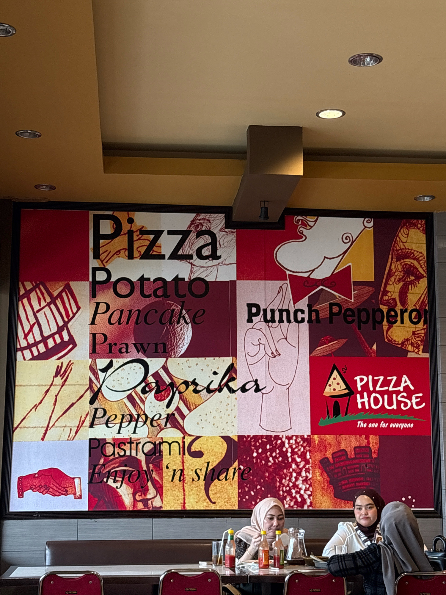 The image captures a brightly lit interior of what appears to be a restaurant. A large, vibrant mural covers the wall behind a booth where two women, both wearing hijabs, are seated at a table. The mural is a collage of various graphic elements and text, with bold black lettering prominently featuring words like "Pizza," "Potato," "Pancake," "Prawn," "Paprika," "Pepper," "Pastrami," and "Enjoy 'n share." Another section of the mural displays "Punch Pepperoni" and a logo for "PIZZA HOUSE, The one for everyone," alongside stylized illustrations of food items, hands, and faces. The women are casually dressed, and one is holding a bottle of sauce. On the table, there are bottles of condiments, glasses, and a pitcher. The lighting suggests overhead fixtures and possibly ambient light from a window. The context of Lhokseumawe, Indonesia, points to a local eatery or a franchise outlet within that city.