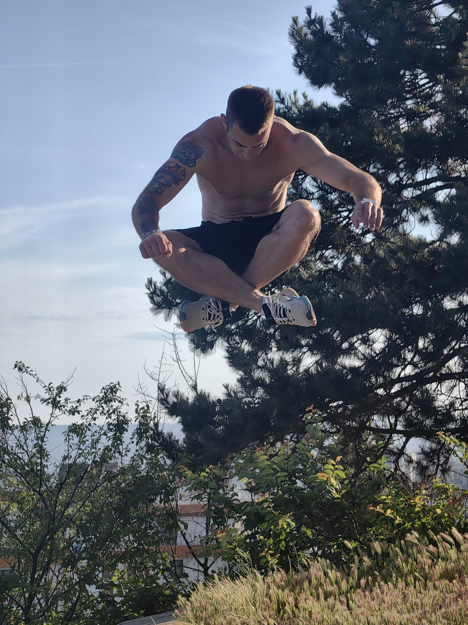 The image shows a shirtless man in mid-air, appearing to be performing a jump or a yoga pose. He is wearing black shorts and athletic shoes. The man has tattoos on his left arm. He is suspended against a clear blue sky with some light clouds. In the background, there is a large pine tree and lush green foliage. The scene appears to be outdoors, possibly in a park or elevated area overlooking a city. The presence of pine trees and the architectural style visible through the foliage suggest a location in Varna, Bulgaria, specifically in an area like the Primorski Park or the Sea Garden, which are known for their extensive greenery and elevated viewpoints. The lighting indicates it is daytime, likely late afternoon due to the warm light.