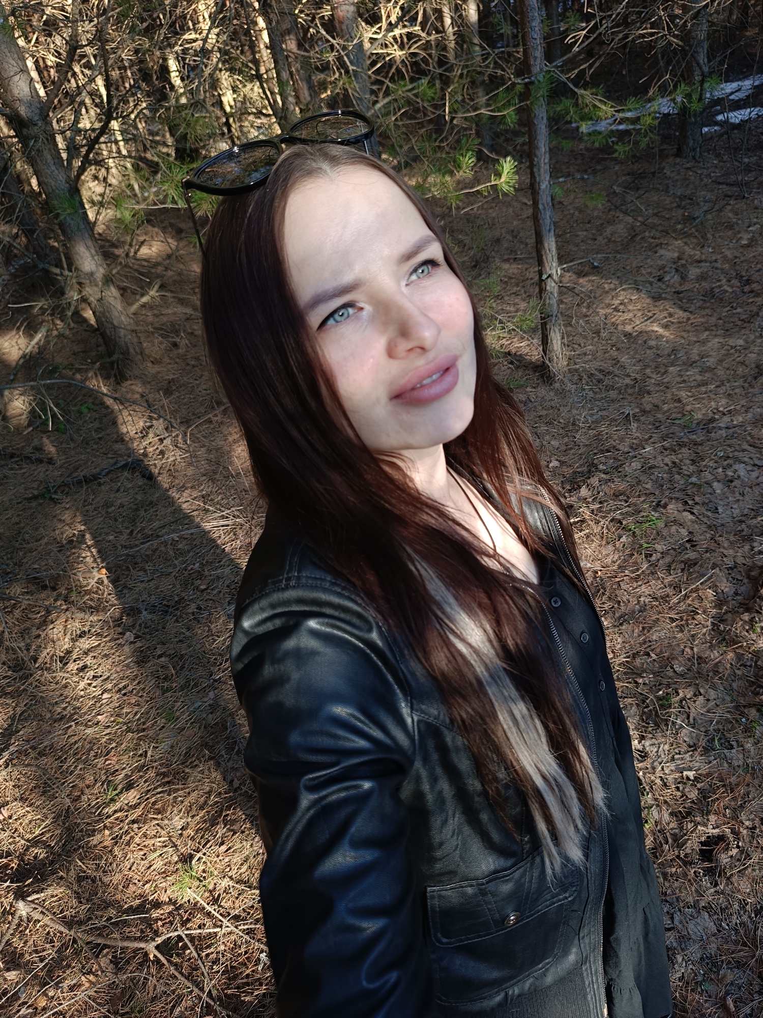 A young woman is captured in a selfie-style shot within a coniferous forest or wooded area, consistent with the landscape of Chaikovsky, Russia. She has long, dark brown hair, with lighter streaks, falling over her shoulders, and sunglasses are perched on top of her head. She is wearing a black leather jacket over a dark shirt. Her gaze is directed upwards and slightly to her right, with a serene and contemplative expression, highlighted by a subtle smile. Her bright blue eyes are a striking feature.
The setting appears to be late winter or early spring, given the bare branches of deciduous trees mingled with conifers, and the ground covered in a thick layer of dried pine needles, twigs, and fallen leaves. One of the cropped hints shows a small patch of snow on the forest floor, reinforcing the seasonal context. The sun is shining brightly, casting long, distinct shadows across the ground, suggesting either early morning or late afternoon. The light illuminates her face from the side, creating a warm glow. There is no visible text in the image.
