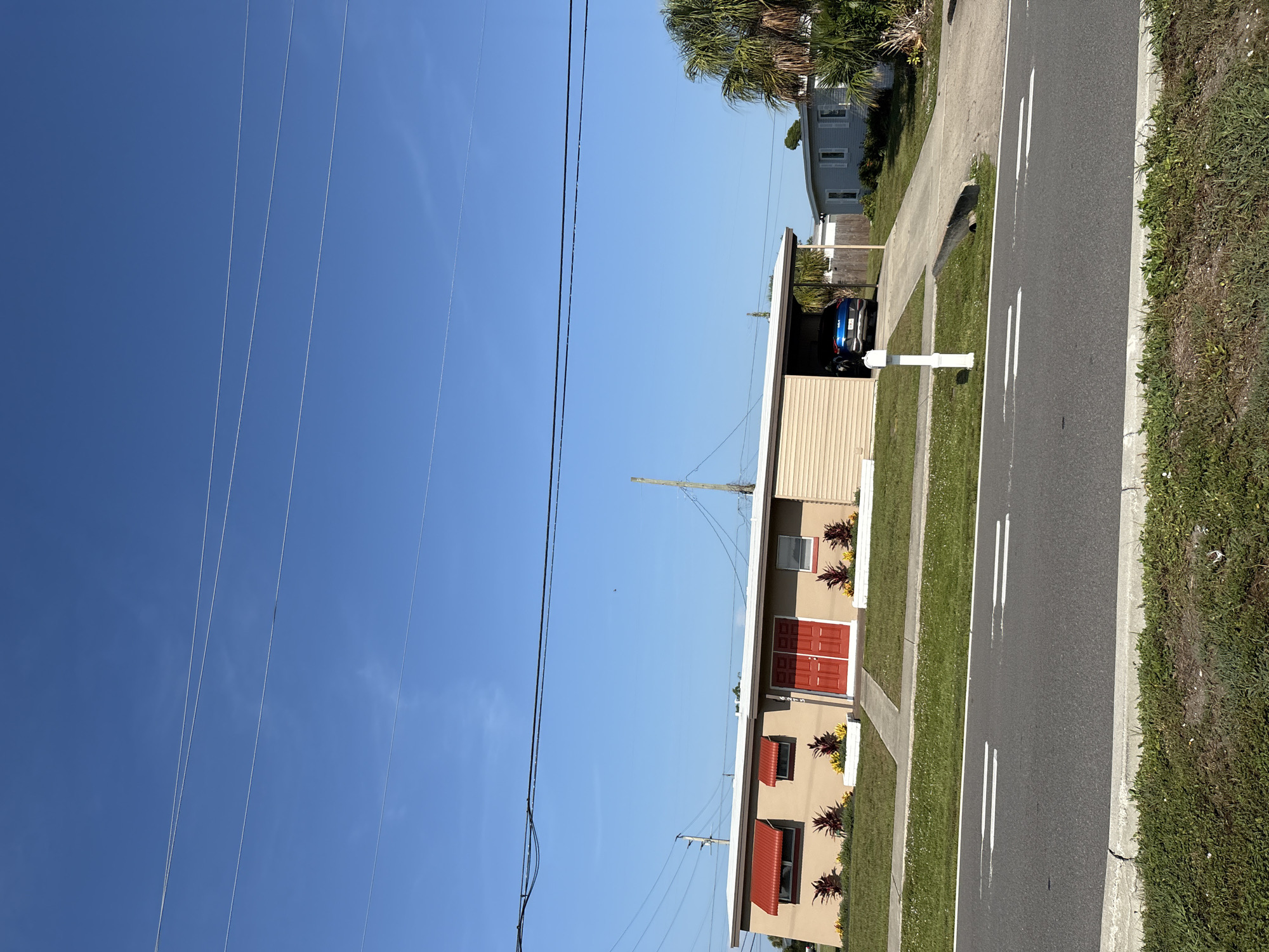 The image depicts a residential street scene on a clear, sunny day. The perspective is from the street, looking towards a beige-colored house with a light brown roof. The house features two prominent red double doors, and two windows with red awnings. Red and yellow plants are visible in planters in front of the house. Adjacent to the house is a covered carport with a blue car parked inside. A white mailbox stands on a post in front of the carport. To the left of the house, a large expanse of clear blue sky is visible with several power lines stretching across it. To the right of the house, a paved road with white lane markings runs alongside a grassy area. In the background, another house with light blue siding and white shutters can be seen, partially obscured by palm trees. The overall scene suggests a quiet suburban or coastal neighborhood in North Port, USA. The time of day appears to be morning or afternoon due to the bright sunlight and distinct shadows. No people are visible in the image. There is no discernible text.