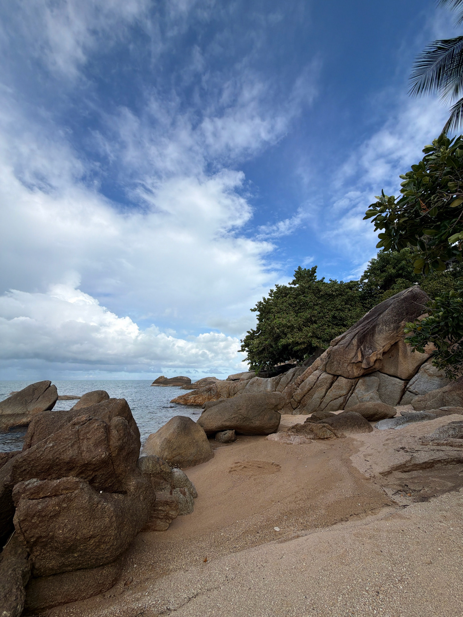 The image depicts a rocky beach on the island of Ko Samui in Thailand. Large, weathered boulders of various sizes are scattered across the sand and in the shallow water. Lush green trees and palm fronds are visible on the right side of the frame, suggesting a tropical environment. The sky above is a vibrant blue with scattered white clouds, indicative of fair weather. The sea is calm, with gentle waves lapping at the shore. The sand appears to be a light brown color, speckled with small shells and pebbles. There are no people or animals visible in the scene. The overall impression is one of a serene and natural coastal landscape.