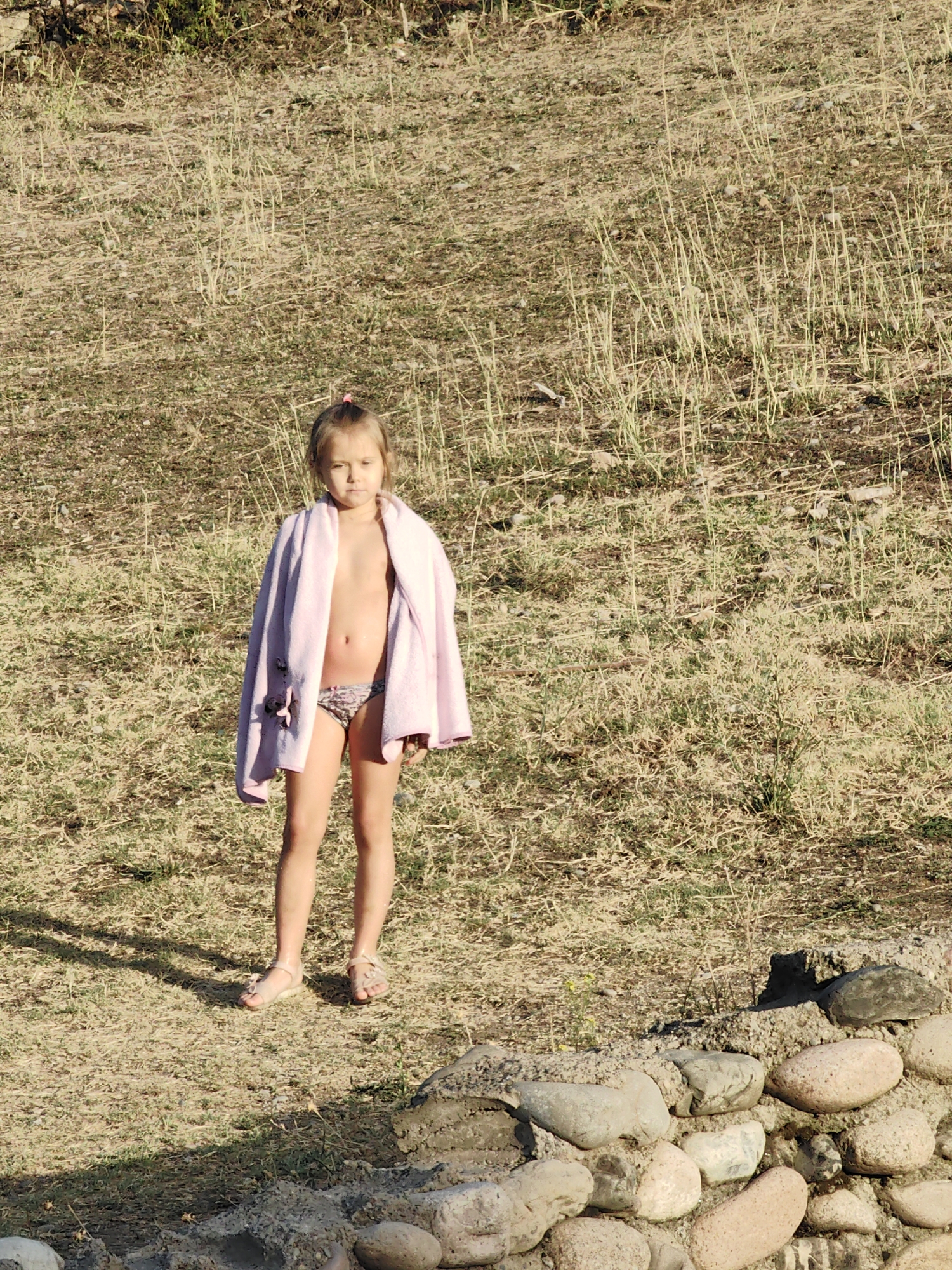 A young girl stands in a field of dry grass and dirt, her body covered by a light pink towel draped over her shoulders. She is wearing patterned bikini bottoms. Her feet are bare except for sandals adorned with small flowers. The ground is uneven and covered with rocks, some of which are stacked in a rough wall at the bottom of the frame. The lighting suggests it is late afternoon, with long shadows cast across the ground. The girl's expression is neutral. The scene appears to be outdoors in a rural area, possibly near a body of water given the presence of a towel. The background is a hillside covered in dry vegetation. The location is identified as Koksayaq Rural District, Kazakhstan. There is no visible text in the image.